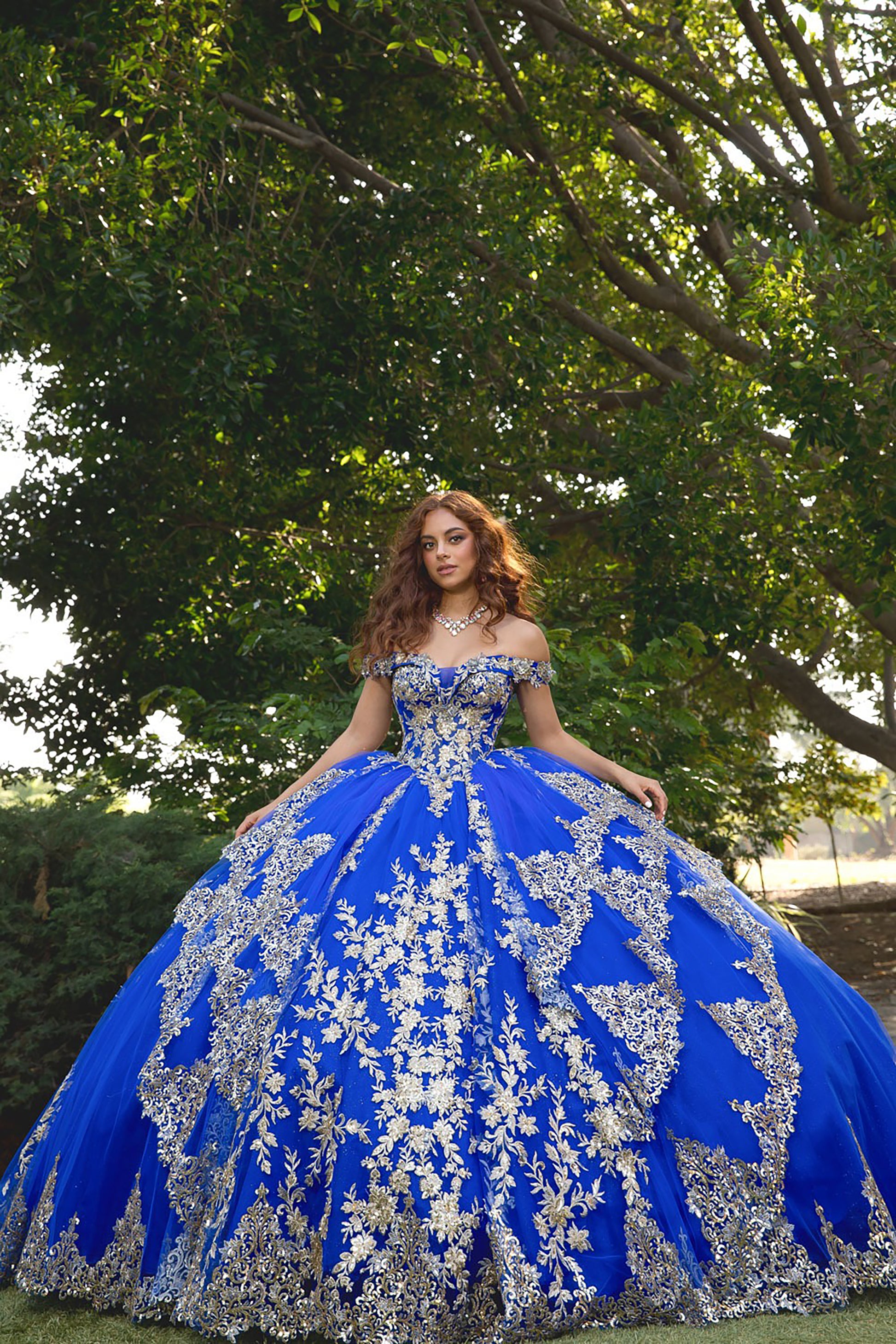 Woman in a blue and silver quinceañera dress standing outdoors with trees in the background