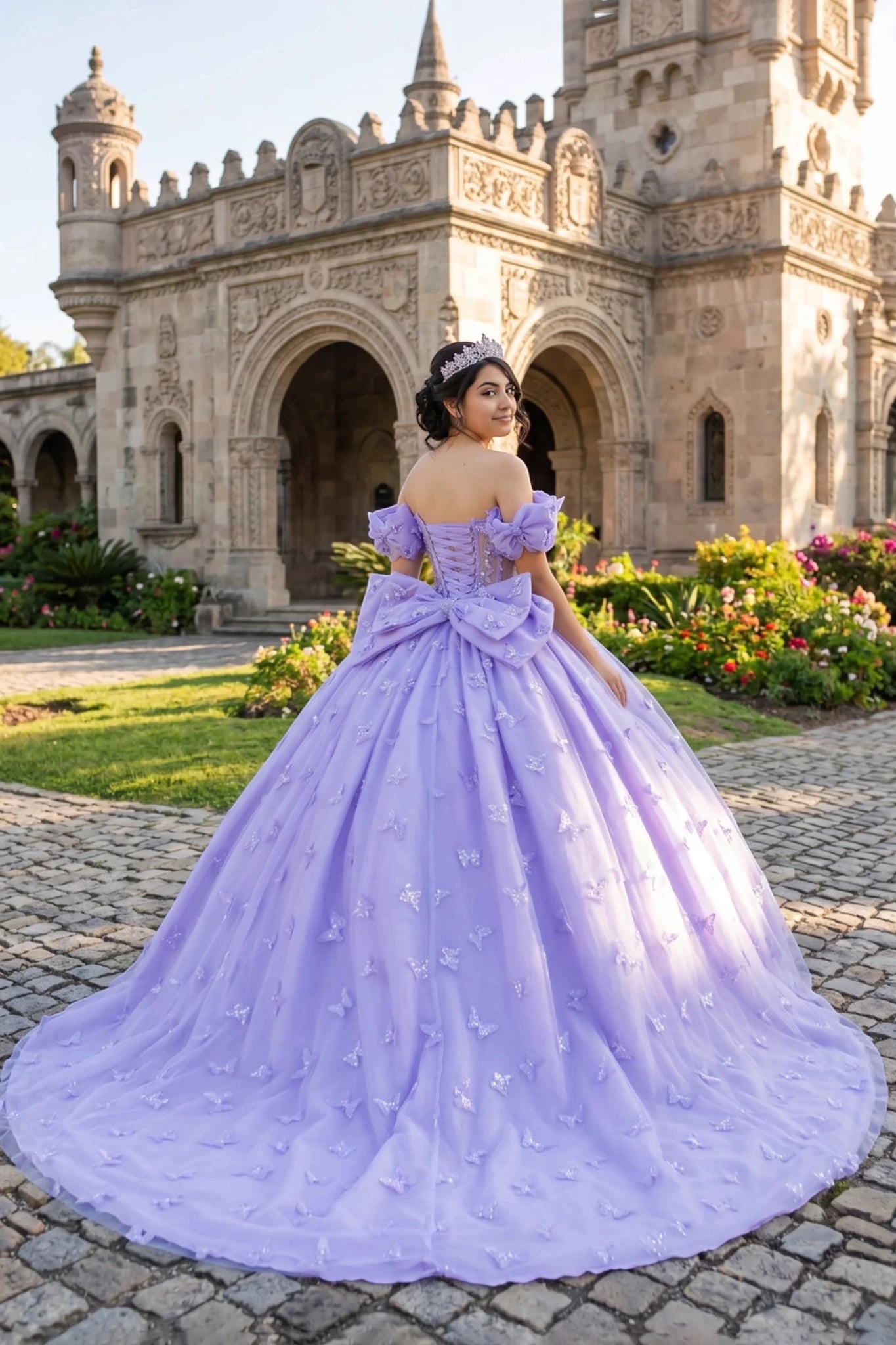 Woman in a lilac quinceanera ball gown with 3D butterflies and  big back bow standing in front of an ornate building with floral gardens.