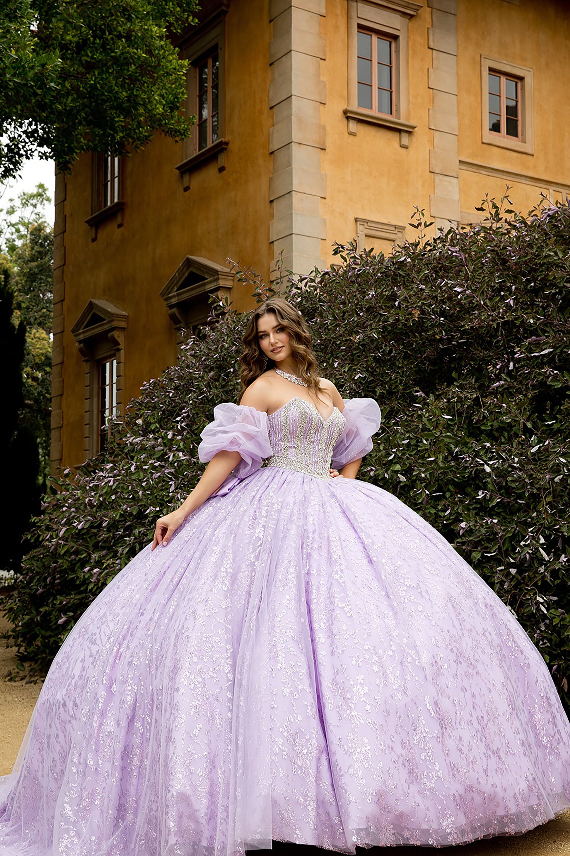 Woman in a lavender ball gown standing in front of a yellow building with greenery.
