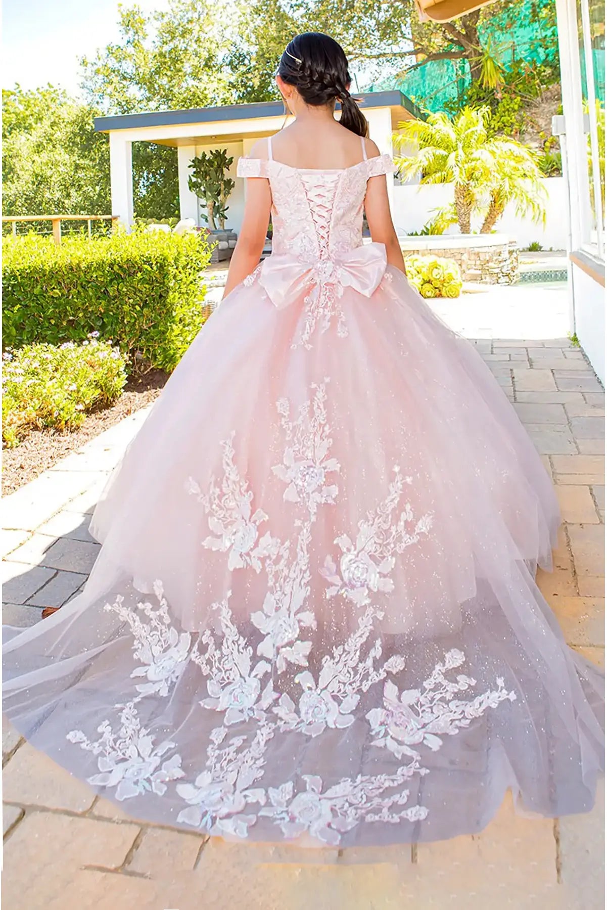Pink ball gown with long train floral lace details and big bow worn by a kid outdoors.