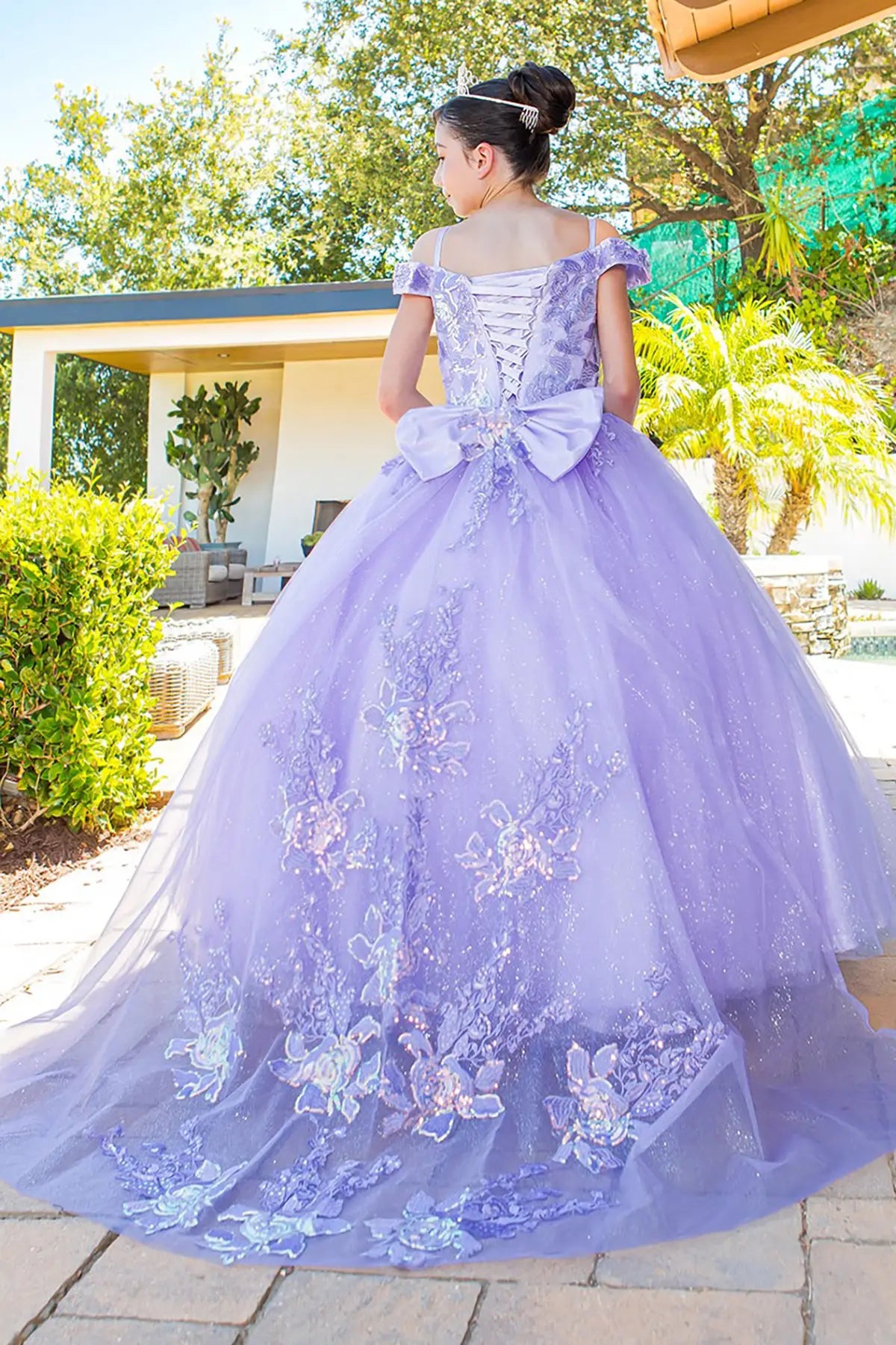 Young girl in a lilac ball gown with 3D appliques and off shoulder standing and showing her back thatis lace up dress with long train outdoors with trees and a pool in the background.