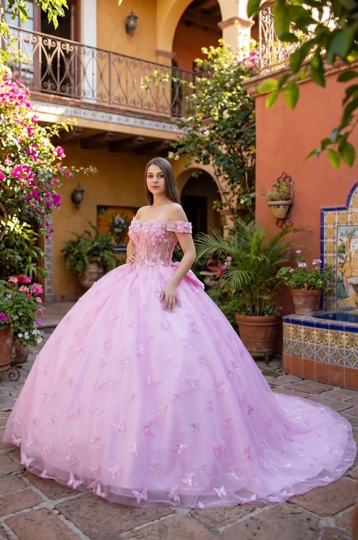 Woman in a pink off-shoulder quinceanera ball gown standing in a decorative courtyard with plants and architecture.