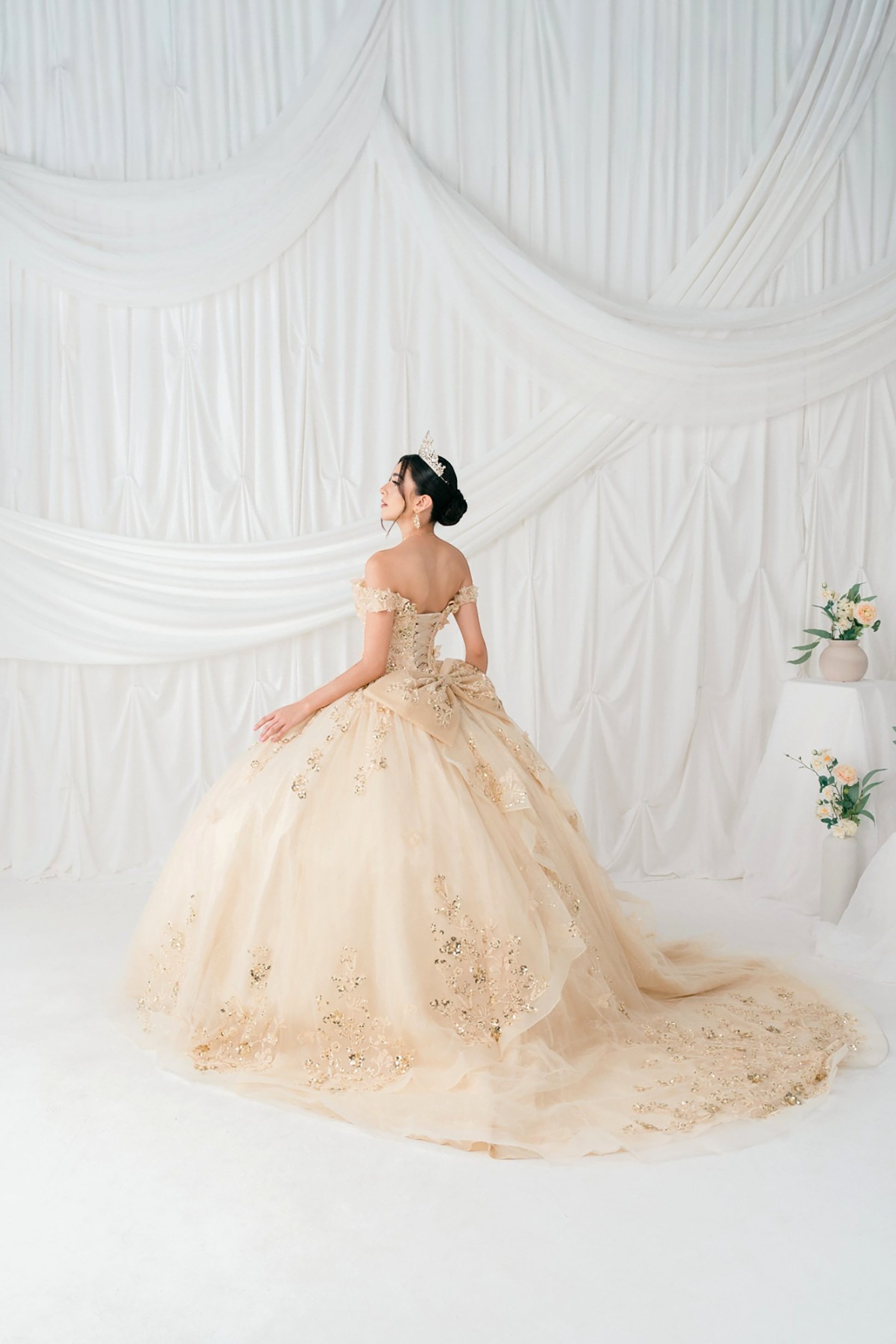 Woman in a champagne dress standing against a white draped curtain backdrop.