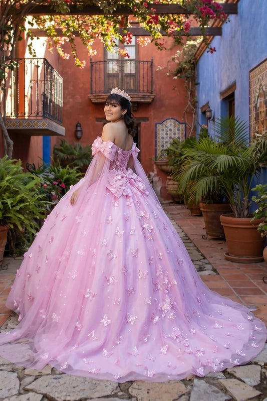 Woman in a pink floral dress standing in an outdoor setting with plants and buildings.