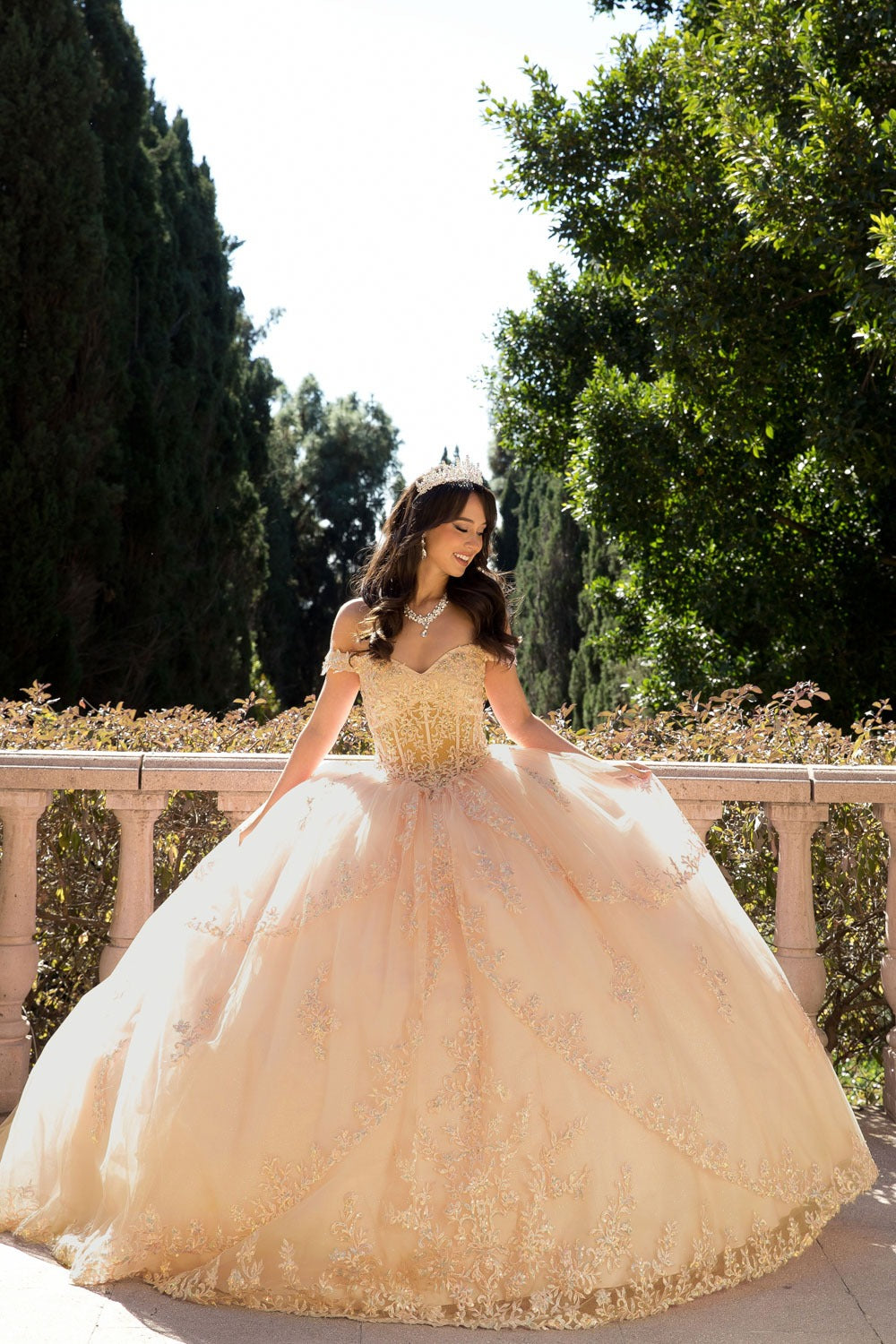 Girl in a champagne quinceanera  standing outdoors with trees in the background