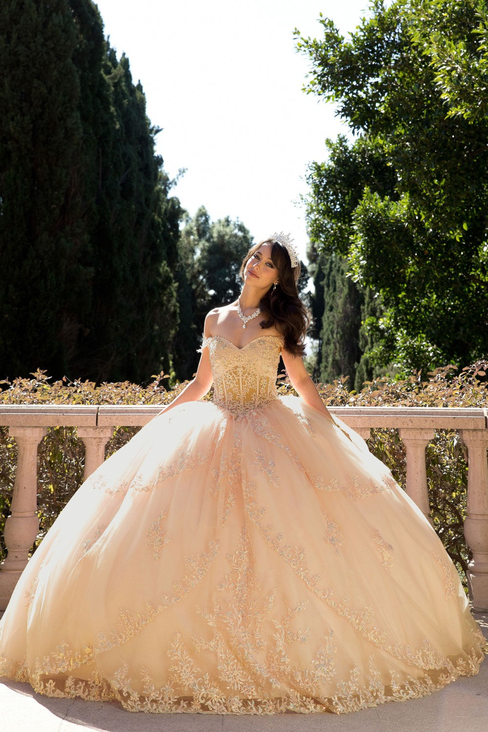 Girl in a champagne quinceanera ball gown standing outdoors with greenery in the background