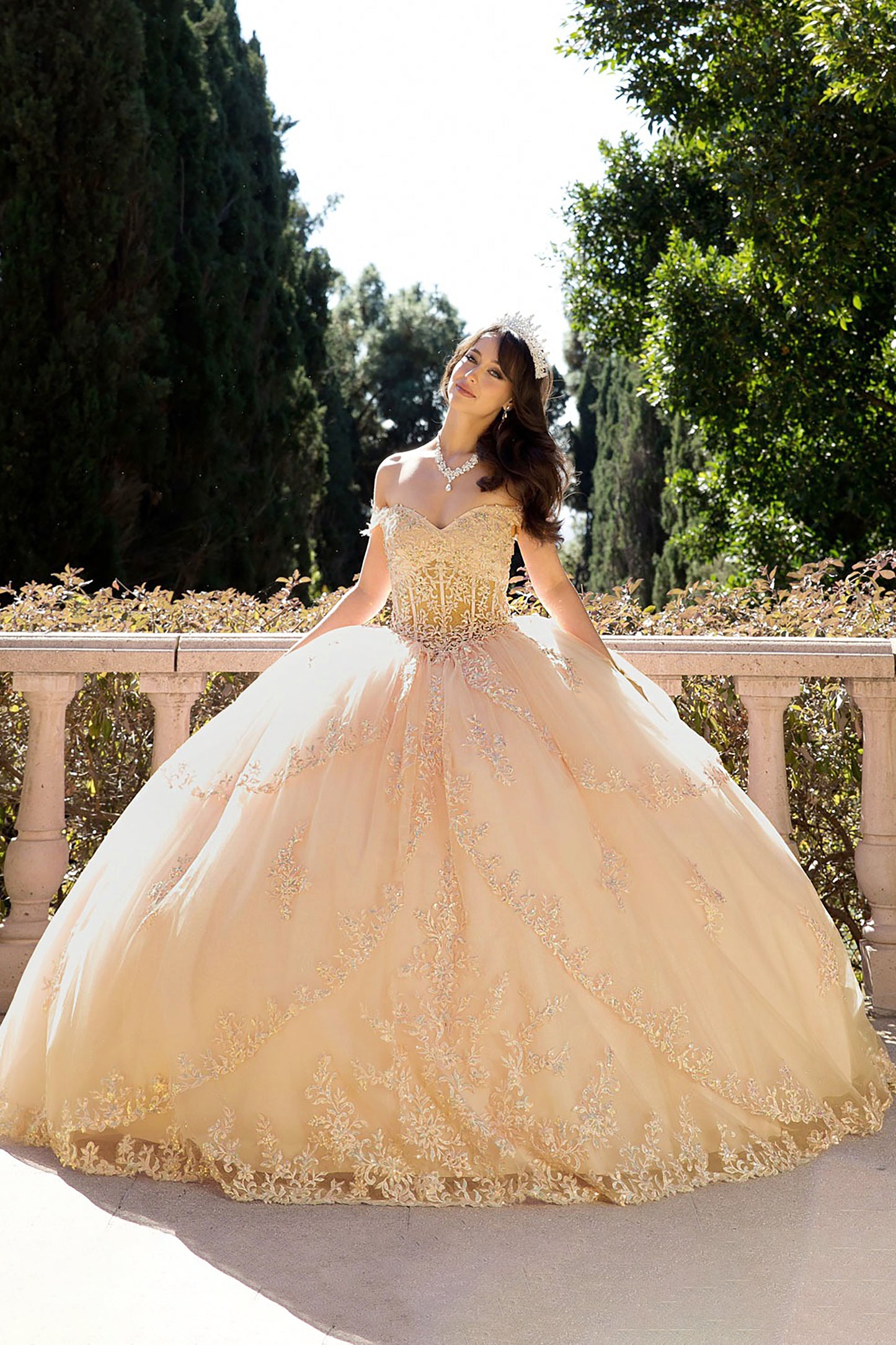 a girl in a champagne ball gown dress standing outdoors with greenery in the background