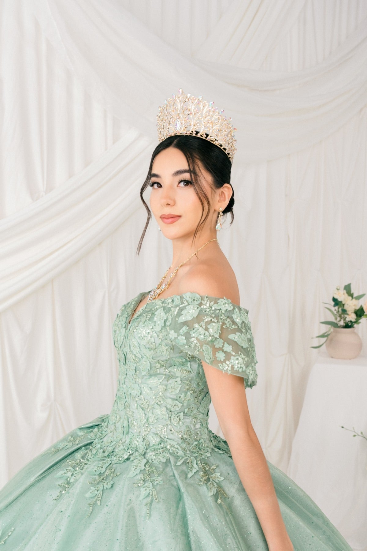 Woman in a light green dress with floral details and a tiara against a white draped curtain background.