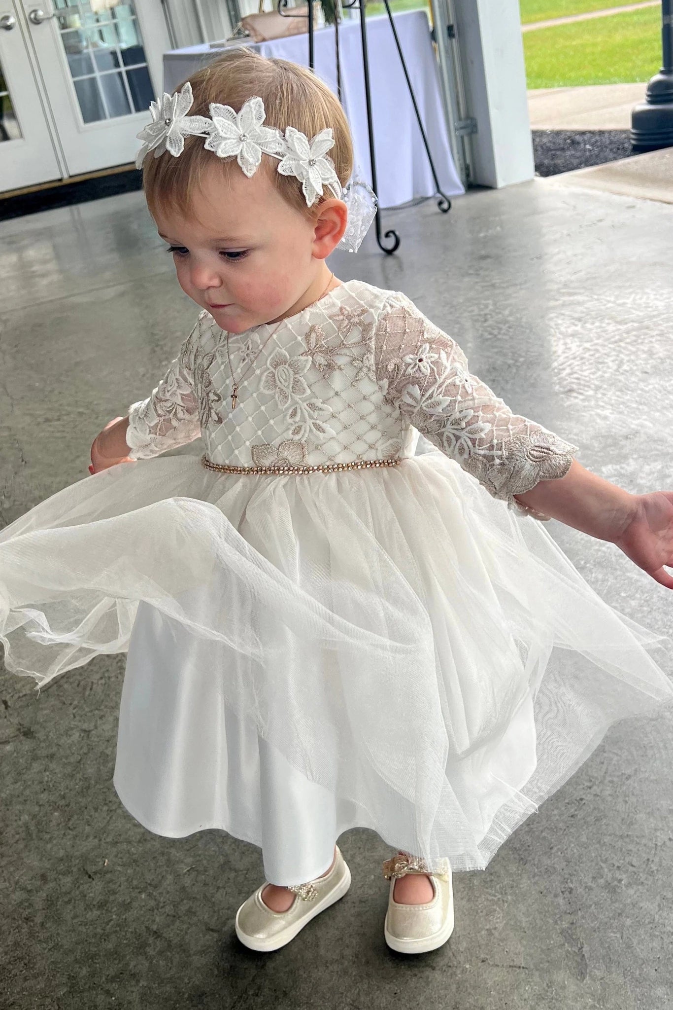 Child in a white lace dress with a floral headband on a concrete floor.