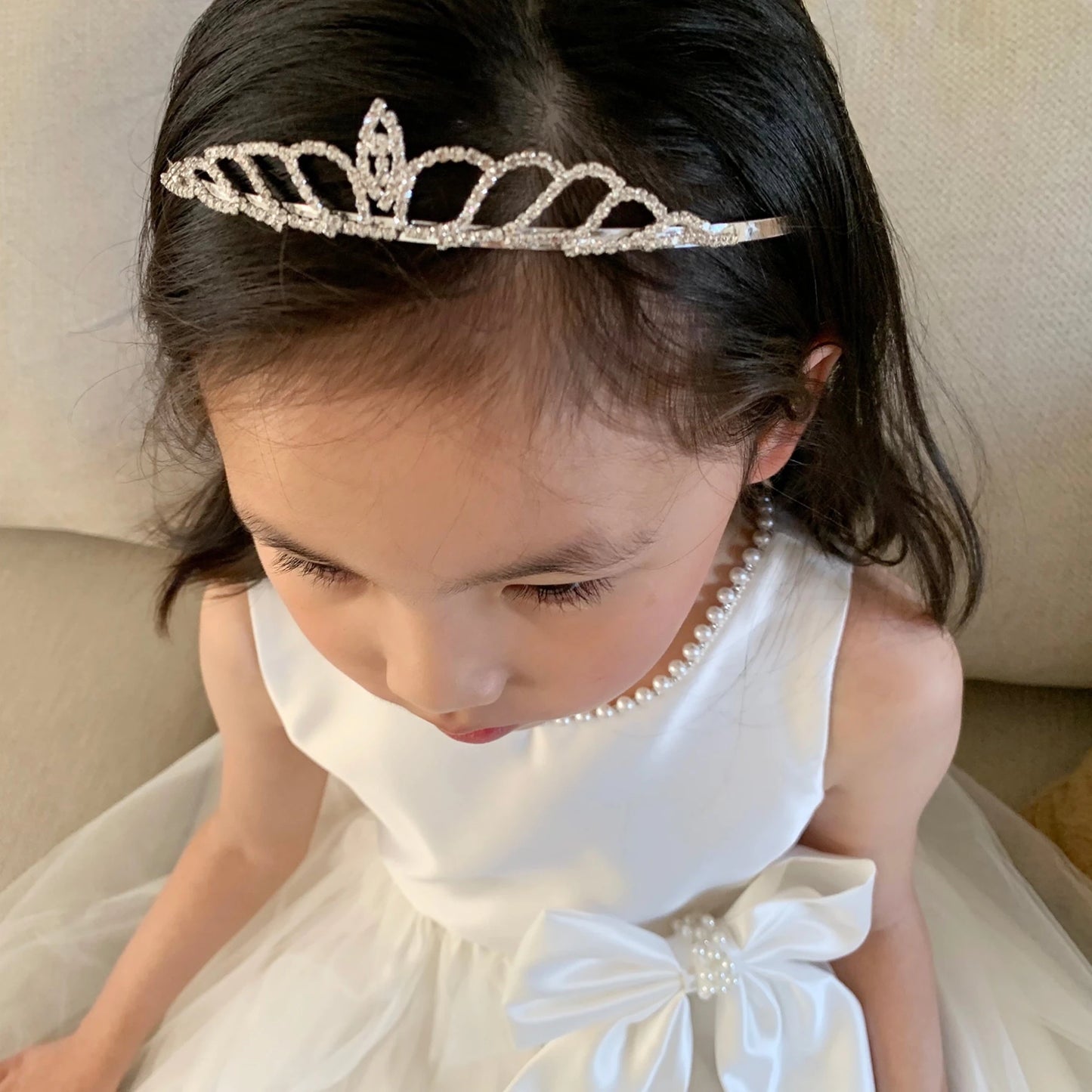 Young girl wearing a Ivory dress and tiara, sitting on a beige couch.