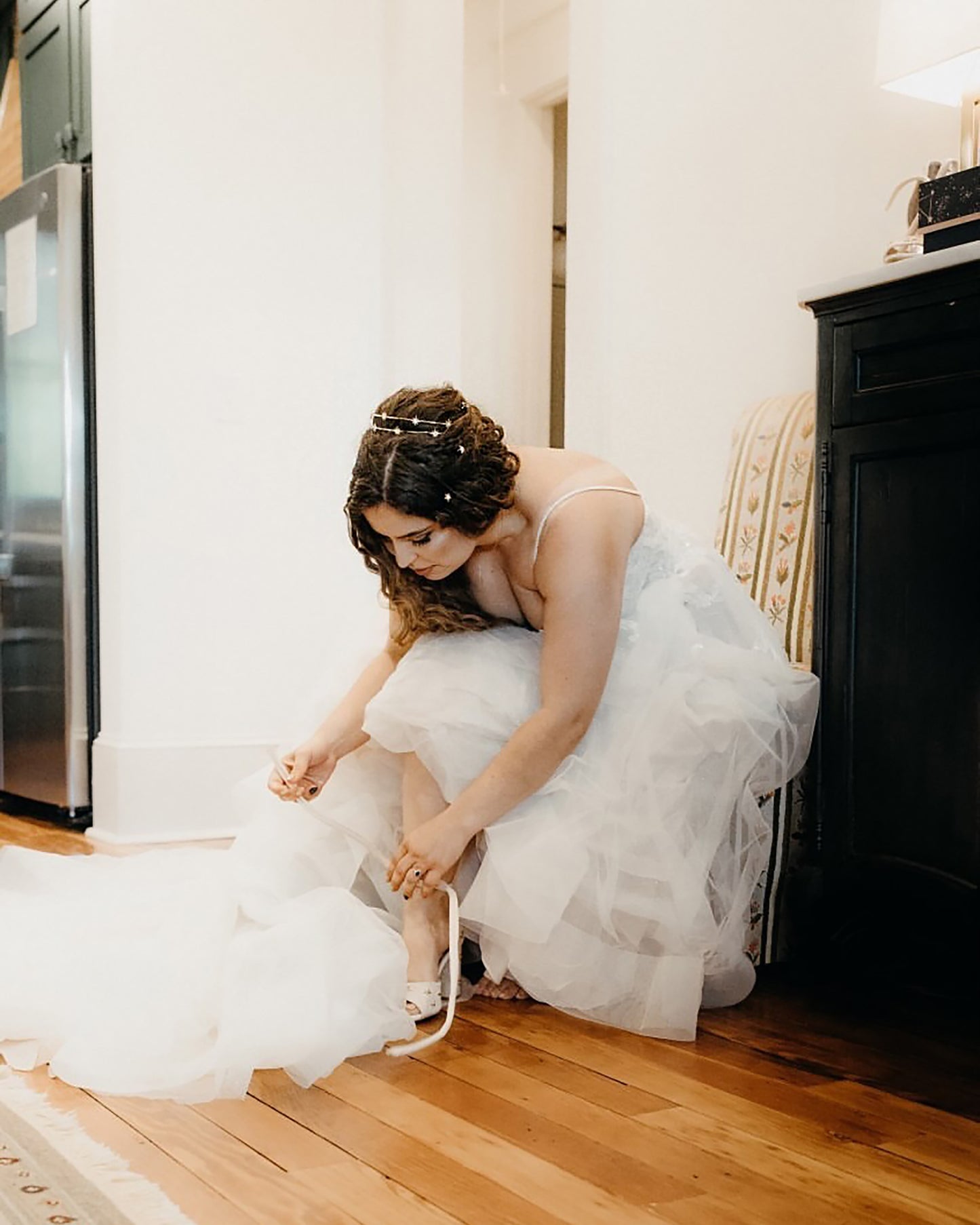 Woman in a white wedding dress and a gold headband adjusting her shoe in a room with wooden floor and black furniture.