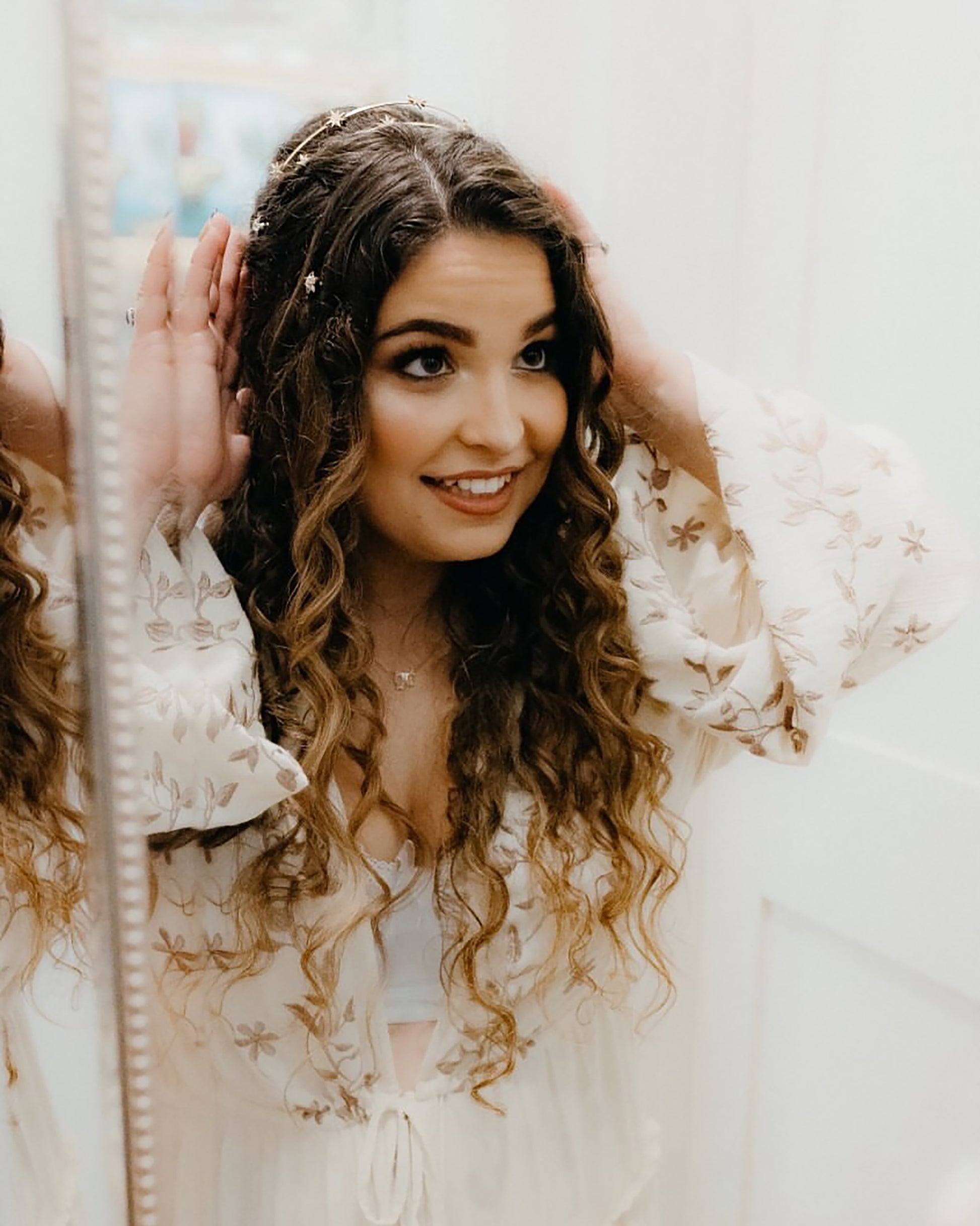 Woman with curly hair wearing a gold rhinestone headband and floral dress in front of a mirror.