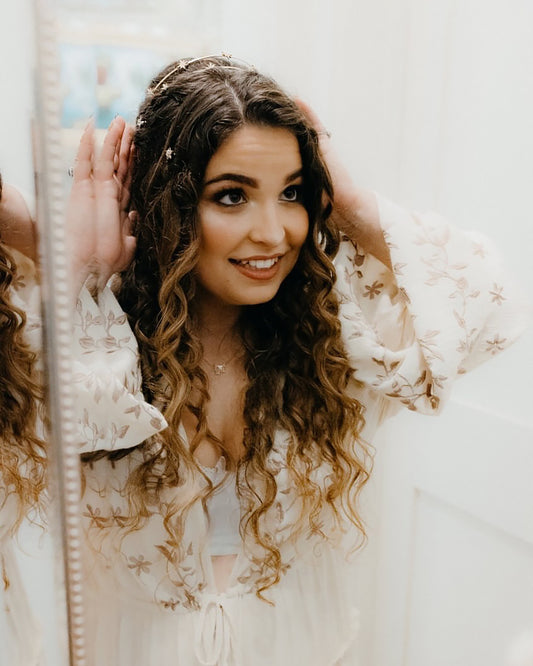 Woman with curly hair wearing a gold rhinestone headband and floral dress in front of a mirror.