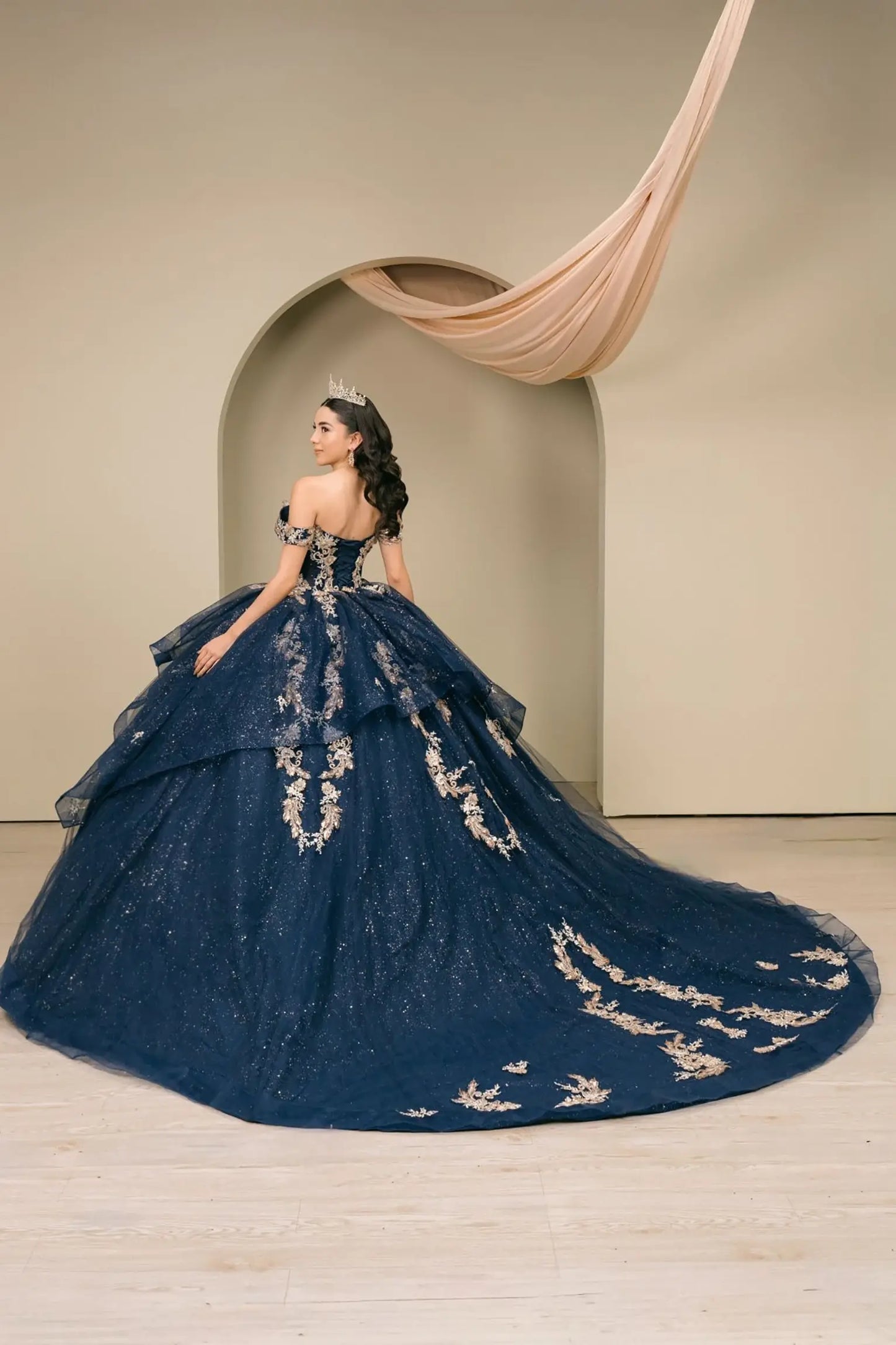 Woman wearing navy quinceañera dress three-quarter view with ornate gold embroidered corset bodice, tiered shimmer tulle skirt, detachable cape, and voluminous train silhouette