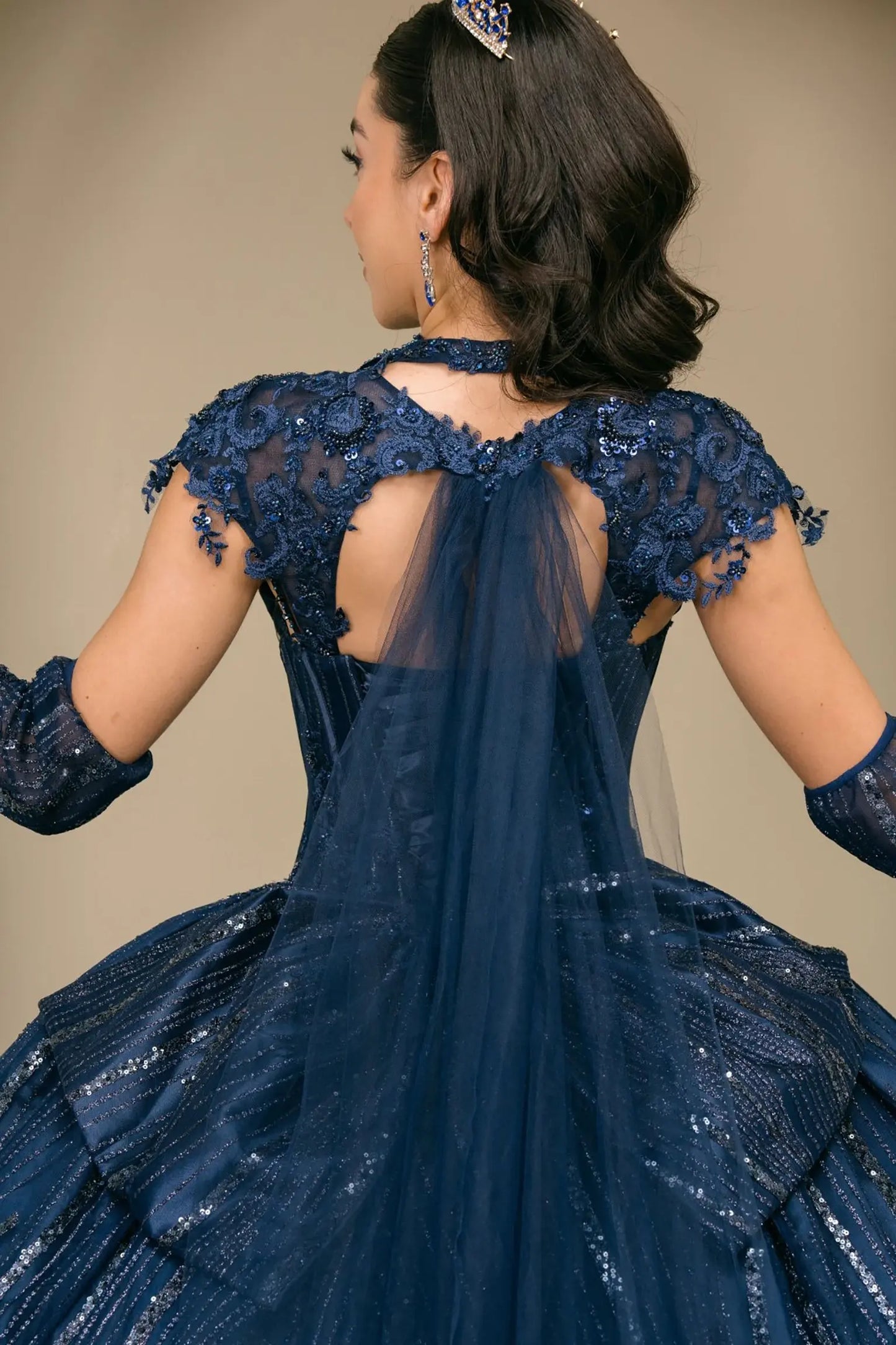 Close-up of woman back-facing in navy quinceañera royal dress with lace-up beaded corset bodice, detachable cape, shimmering textured skirt, cathedral train, and gloves