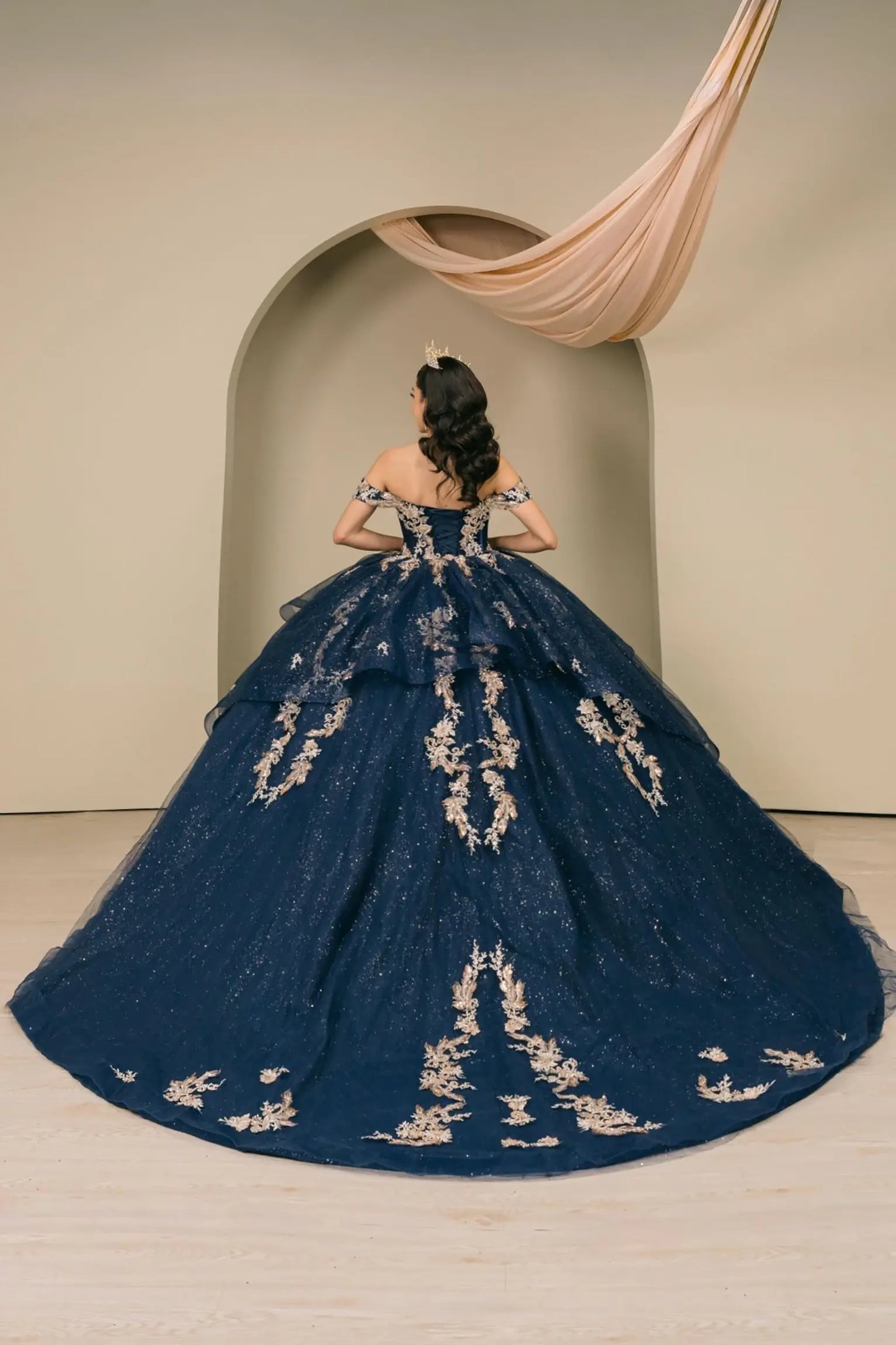 Woman back-facing in navy quinceañera dress with lace-up corset bodice, gold embroidery, detachable cape layer, and dramatic extended train