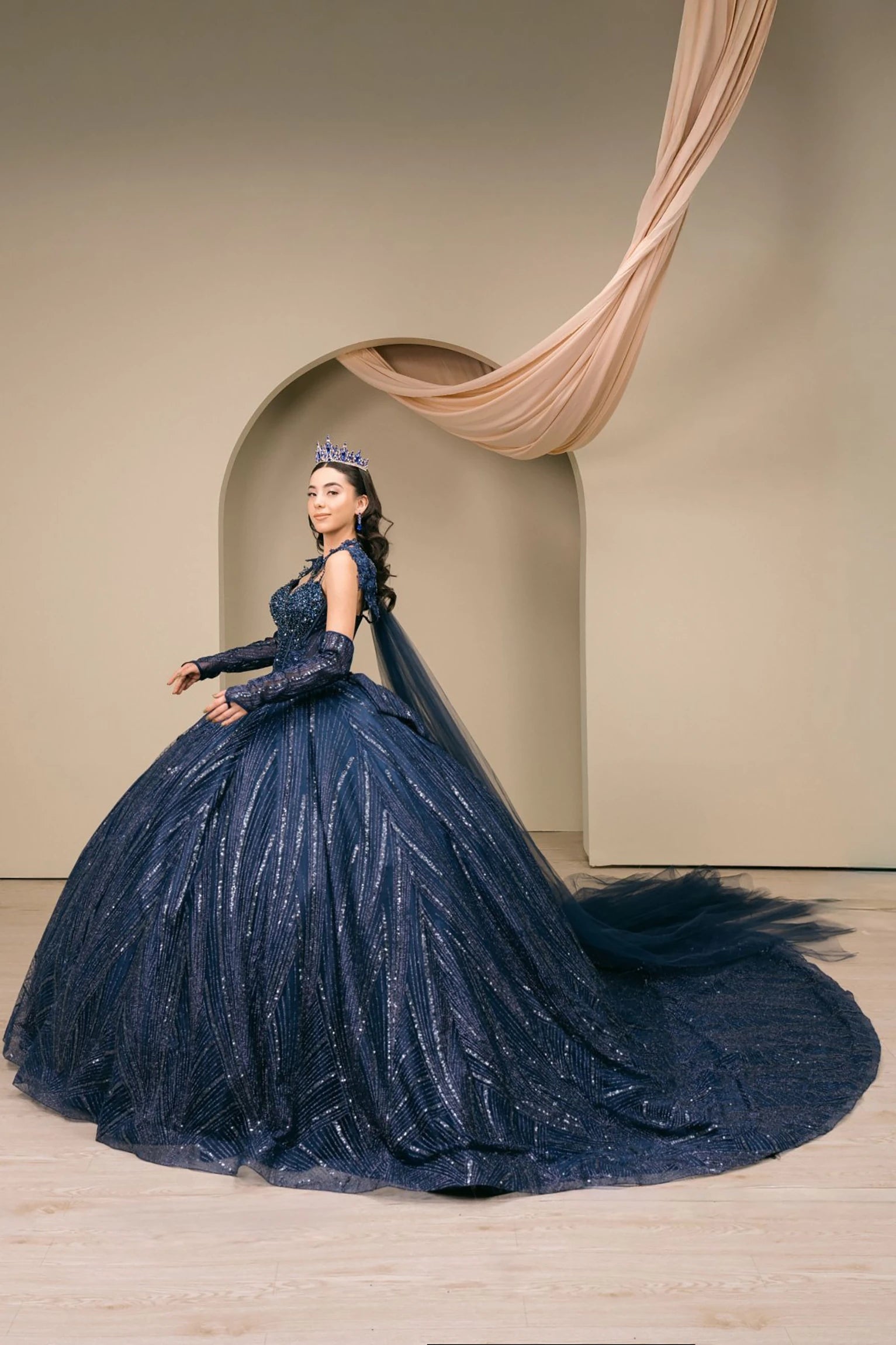Woman wearing navy quinceañera royal dress side-facing with beaded corset bodice, shimmering textured skirt, cathedral train, detachable cape, choker neckline, and gloves