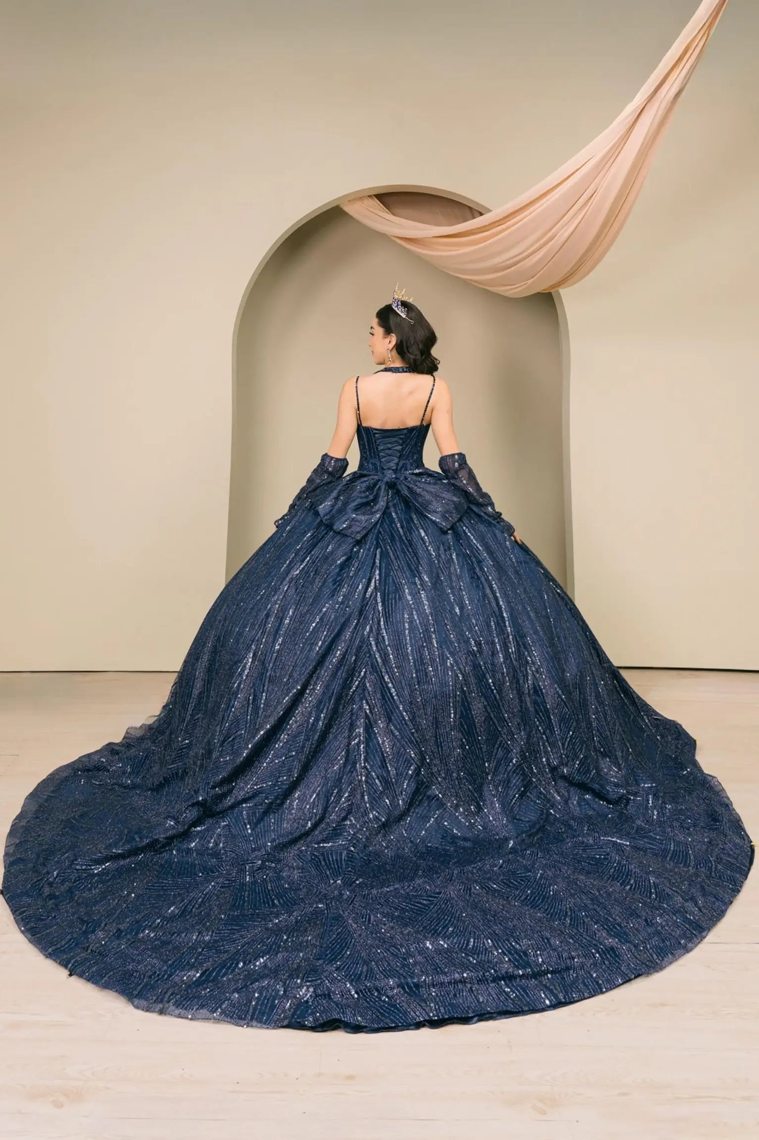 Woman back-facing in navy quinceañera royal dress with lace-up beaded corset bodice, shimmering textured skirt, cathedral train, detachable cape, and gloves