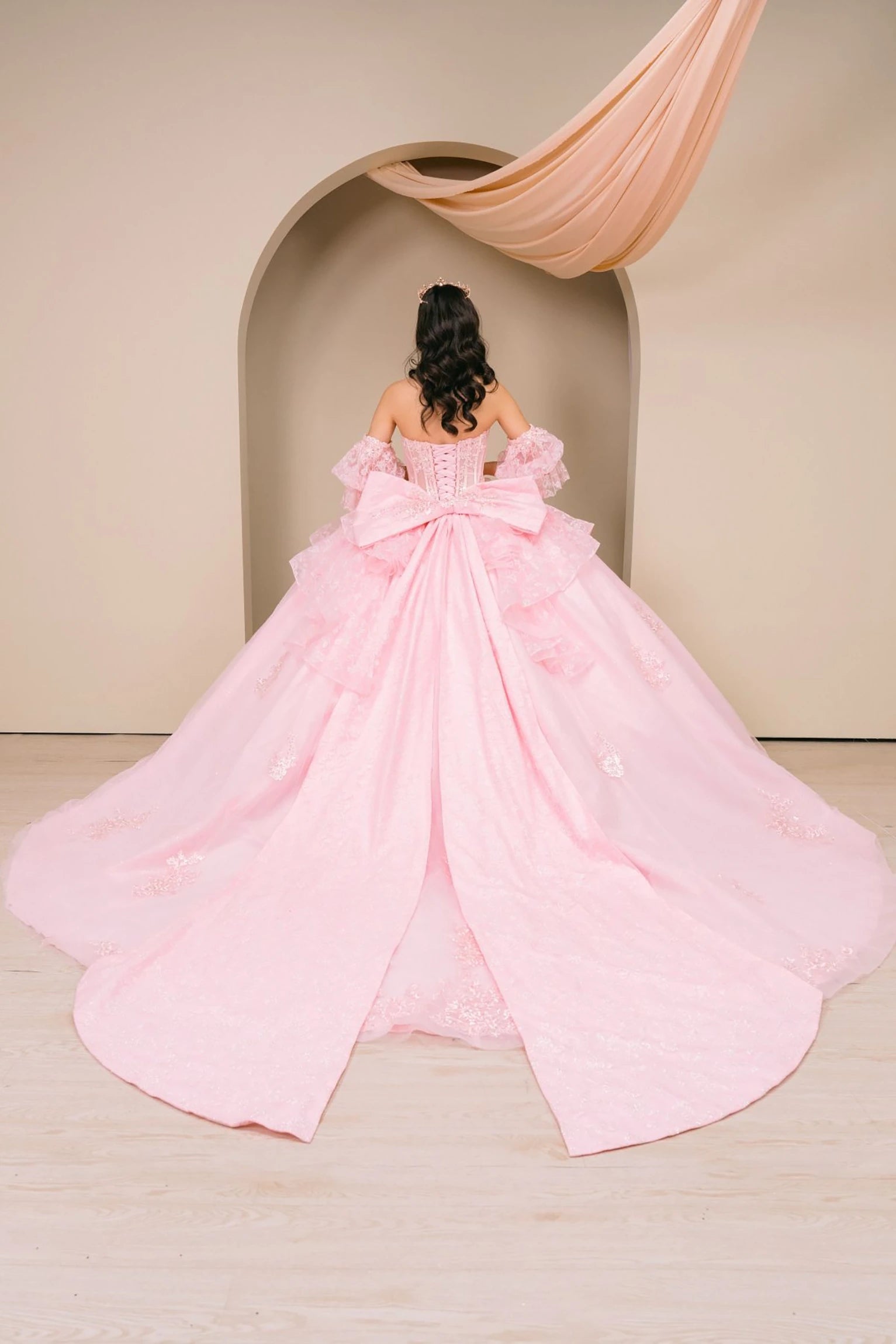 Woman back-facing in pink off-shoulder sweetheart quinceañera dress with lace-up corset bodice, beaded embroidery, and cascading layered tulle organza skirt