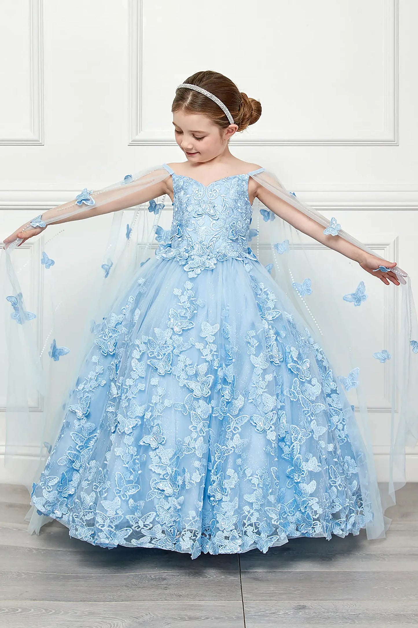 Young girl standing front-facing in blue kids mini quinceañera dress with off-shoulder 3D butterfly floral bodice, layered tulle skirt, and detachable cape
