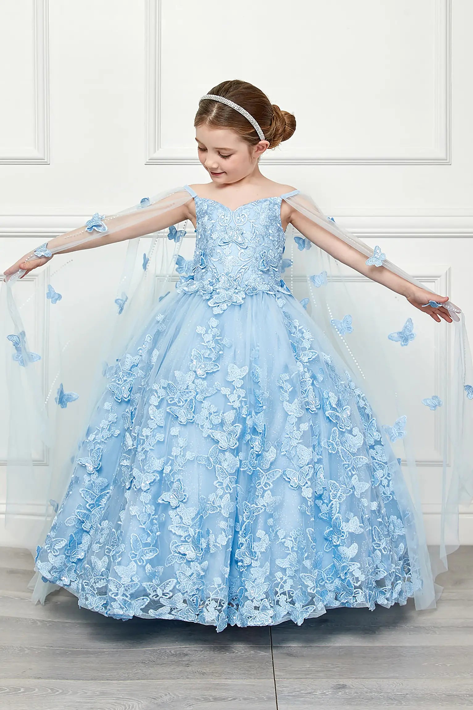 Young girl standing front-facing in blue kids mini quinceañera dress with off-shoulder 3D butterfly floral bodice, layered tulle skirt, and detachable cape