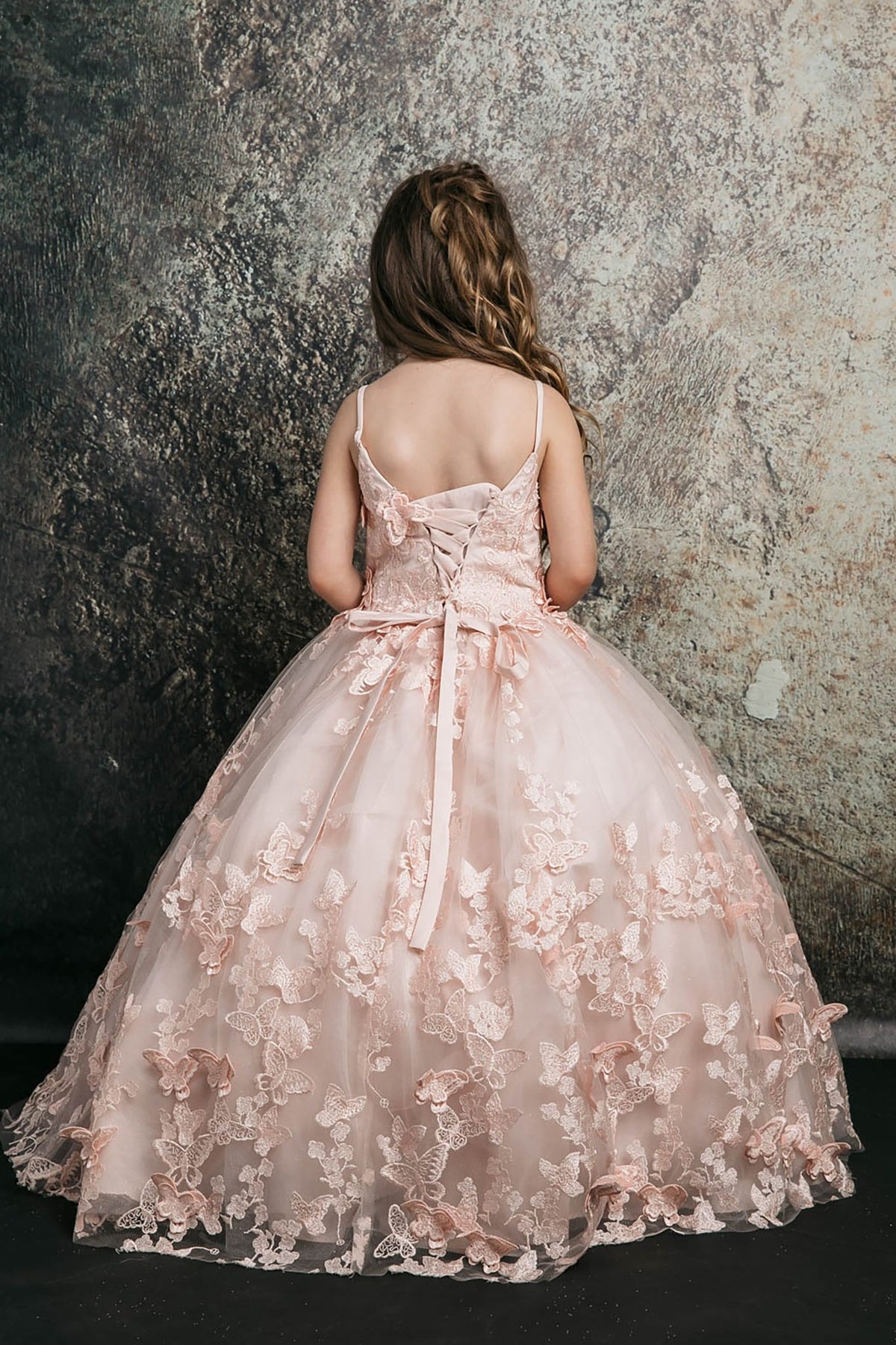 Back view of a blush kids mini quinceanera mini quince dress with lace-up corset, oversized bow, butterfly floral appliques, and detachable cape.