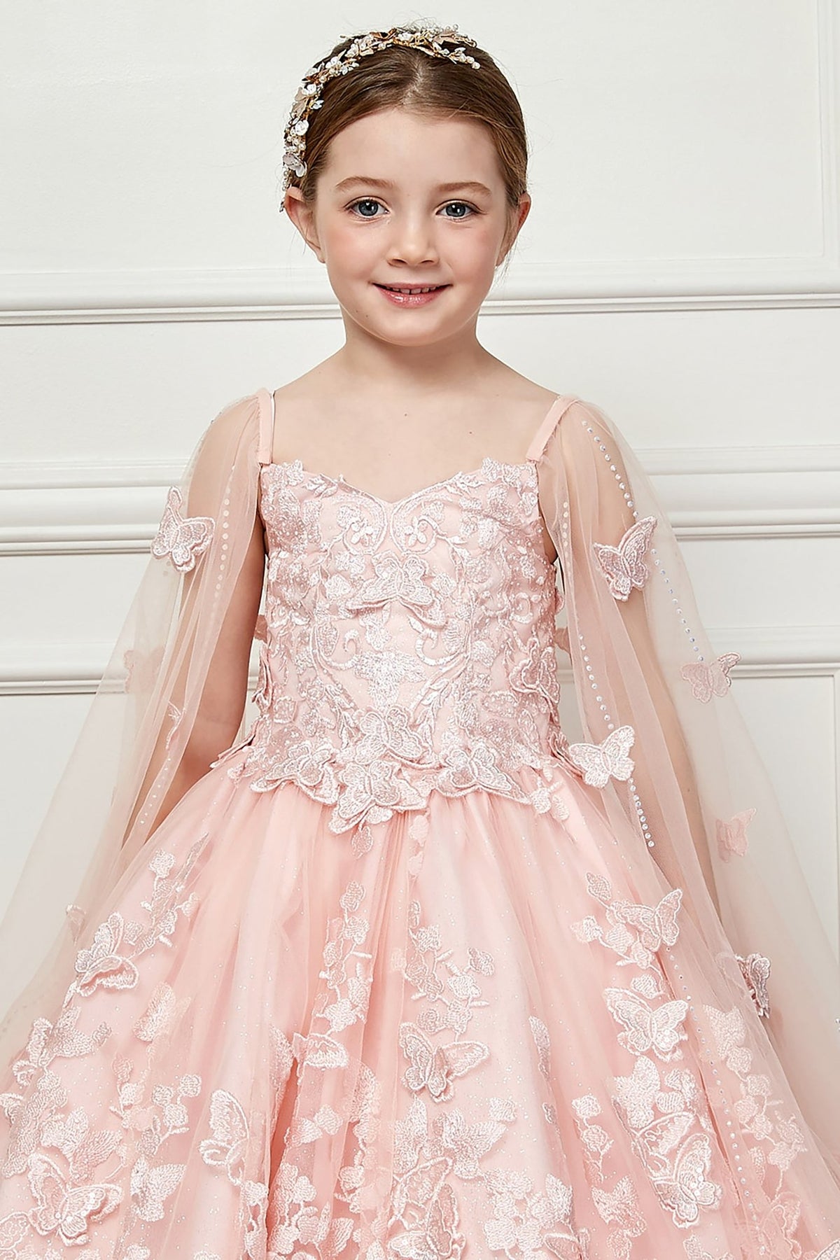 Zoomed front view of a blush kids mini quinceanera mini quince dress highlighting butterfly floral appliques, gem accents, and glitter tulle skirt.