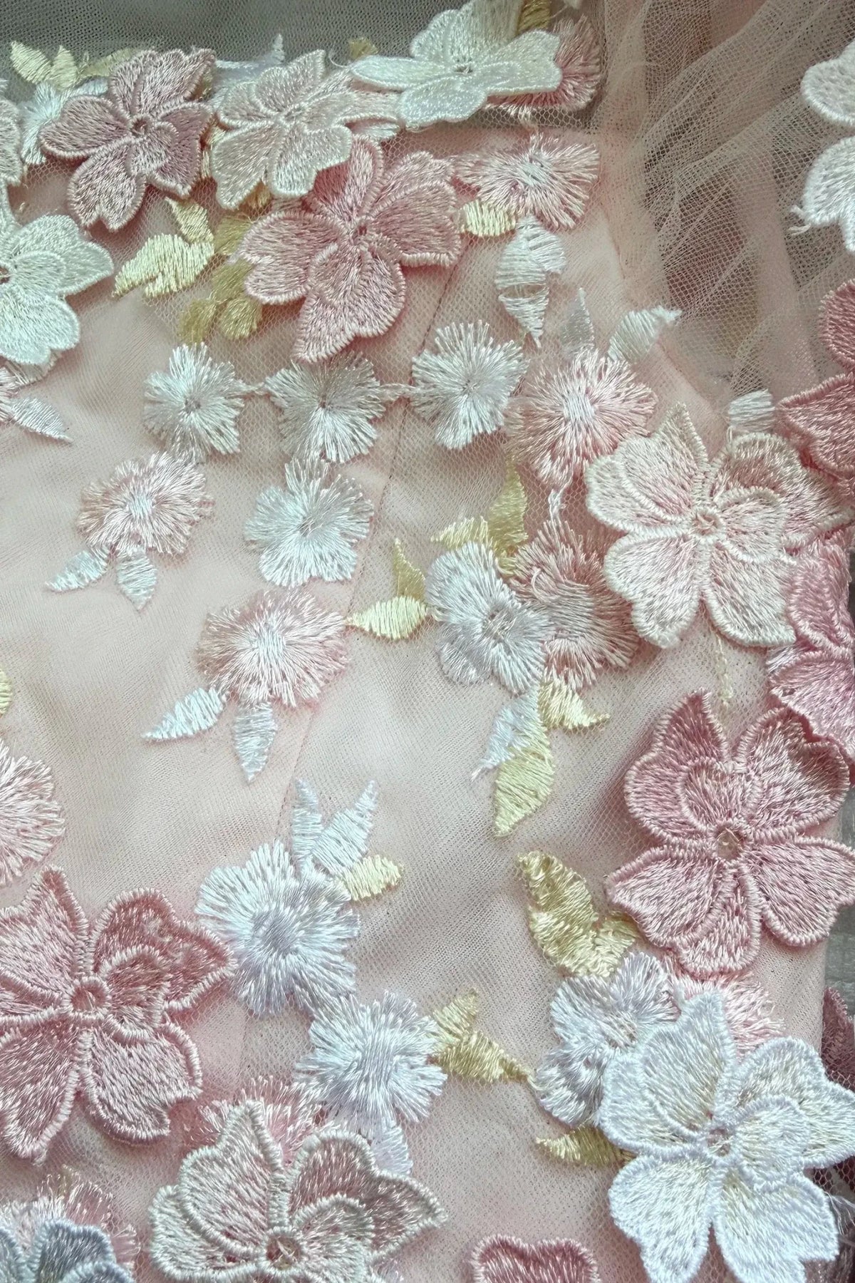 Close-up of a light blush pink embroidered fabric with layered floral applique in pink, white, and pale gold tones.