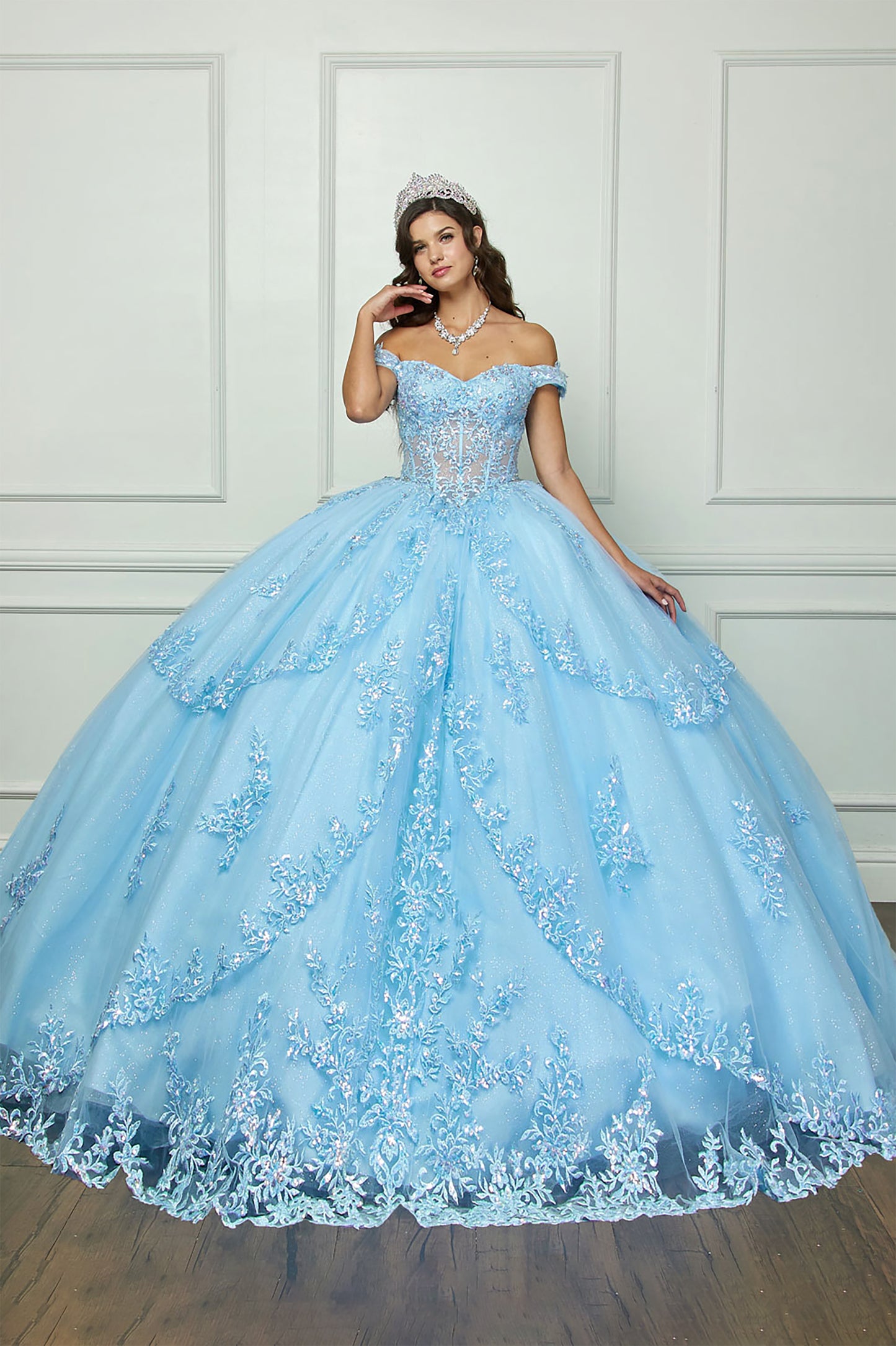 girl in a light blue ball gown with floral details against a white paneled wall.