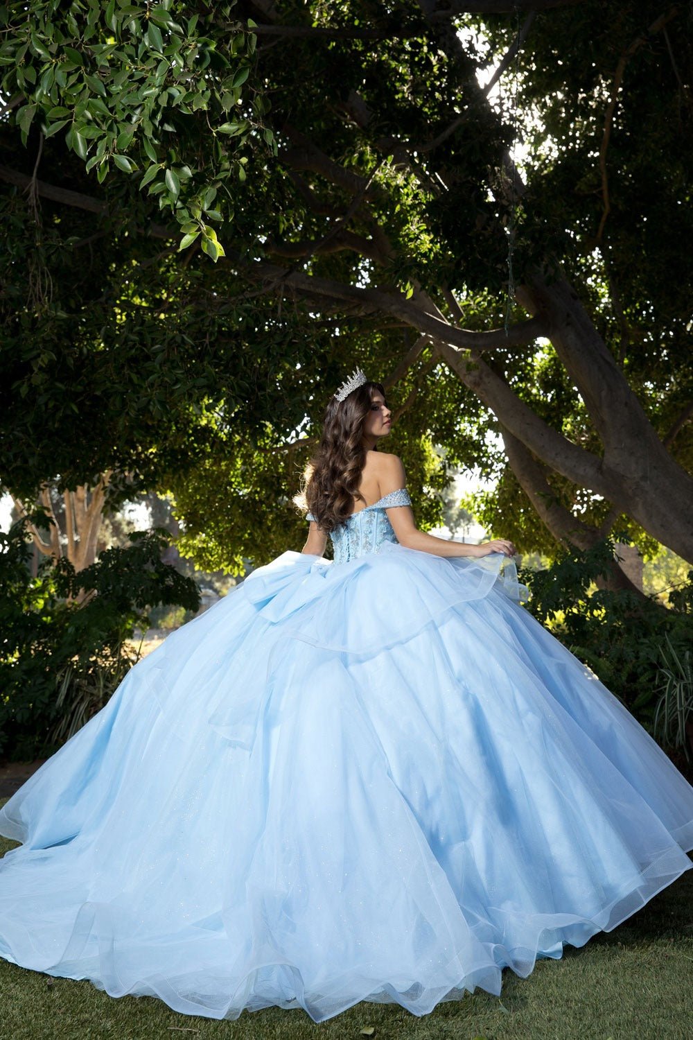 Woman in a light blue quince ball gown showing her back with big large bow standing outdoors with trees in the background