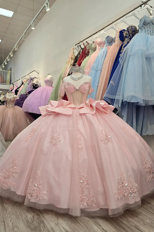 Elegant full-length blush quinceañera ball gown on a mannequin, featuring a beaded sweetheart corset bodice, sparkling silver tiara, and a voluminous glitter tulle skirt adorned with intricate floral lace appliqués and tiered satin ruffles in a boutique showroom.