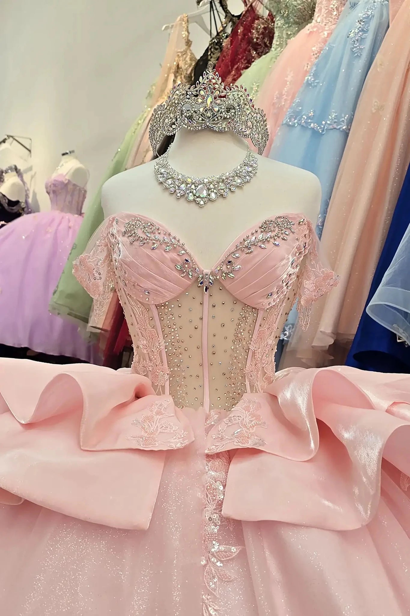 Elegant blush quinceañera dress with a beaded sweetheart corset bodice and sparkling silver crown on a mannequin, featuring intricate embroidery and a voluminous ruffled satin skirt in a bridal boutique setting.