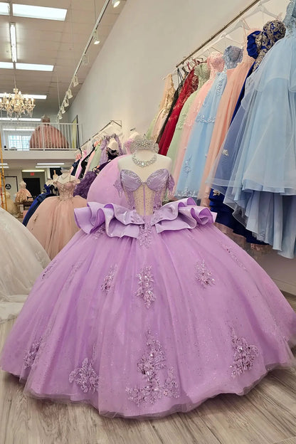 Elegant full-length liliac quinceañera ball gowns on mannequins, featuring beaded sweetheart corset bodices with sheer panels, off-the-shoulder draped sleeves, and adjustable lace-up backs. The ensemble includes sparkling silver tiaras and matching rhinestone necklaces. The gowns showcase voluminous glitter tulle skirts adorned with intricate floral lace appliqués and tiered, sculptural satin ruffles, displayed in a boutique showroom.