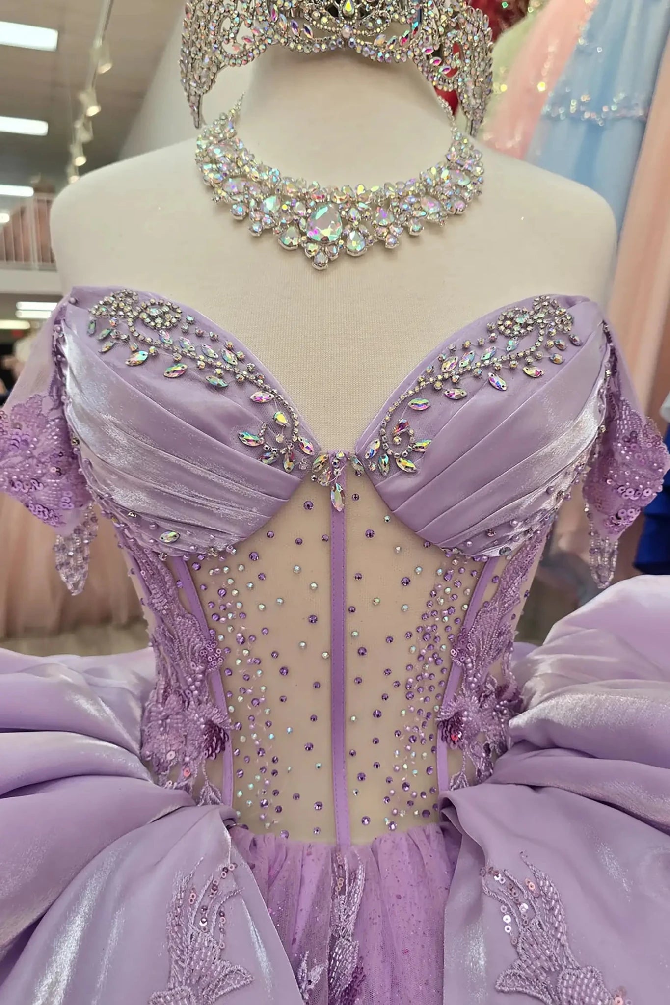 Lilac quinceañera ball gowns on mannequins, featuring beaded sweetheart corset bodices, sparkling silver tiaras, and matching rhinestone necklaces. The gowns showcase voluminous glitter tulle skirts adorned with intricate floral lace appliqués and tiered satin ruffles, displayed in a boutique showroom.