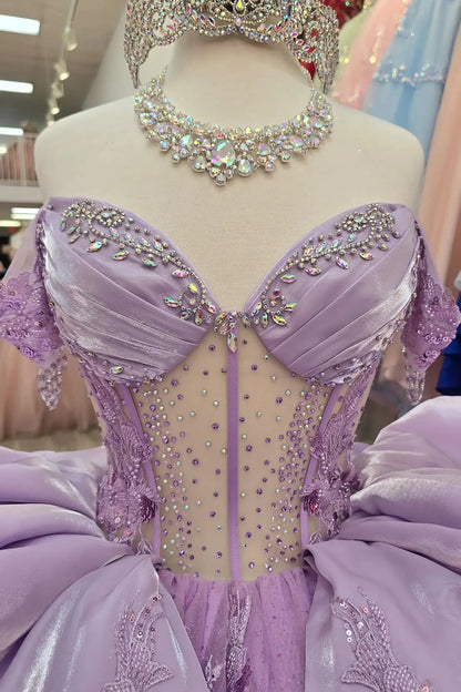 Lilac quinceañera ball gowns on mannequins, featuring beaded sweetheart corset bodices, sparkling silver tiaras, and matching rhinestone necklaces. The gowns showcase voluminous glitter tulle skirts adorned with intricate floral lace appliqués and tiered satin ruffles, displayed in a boutique showroom.