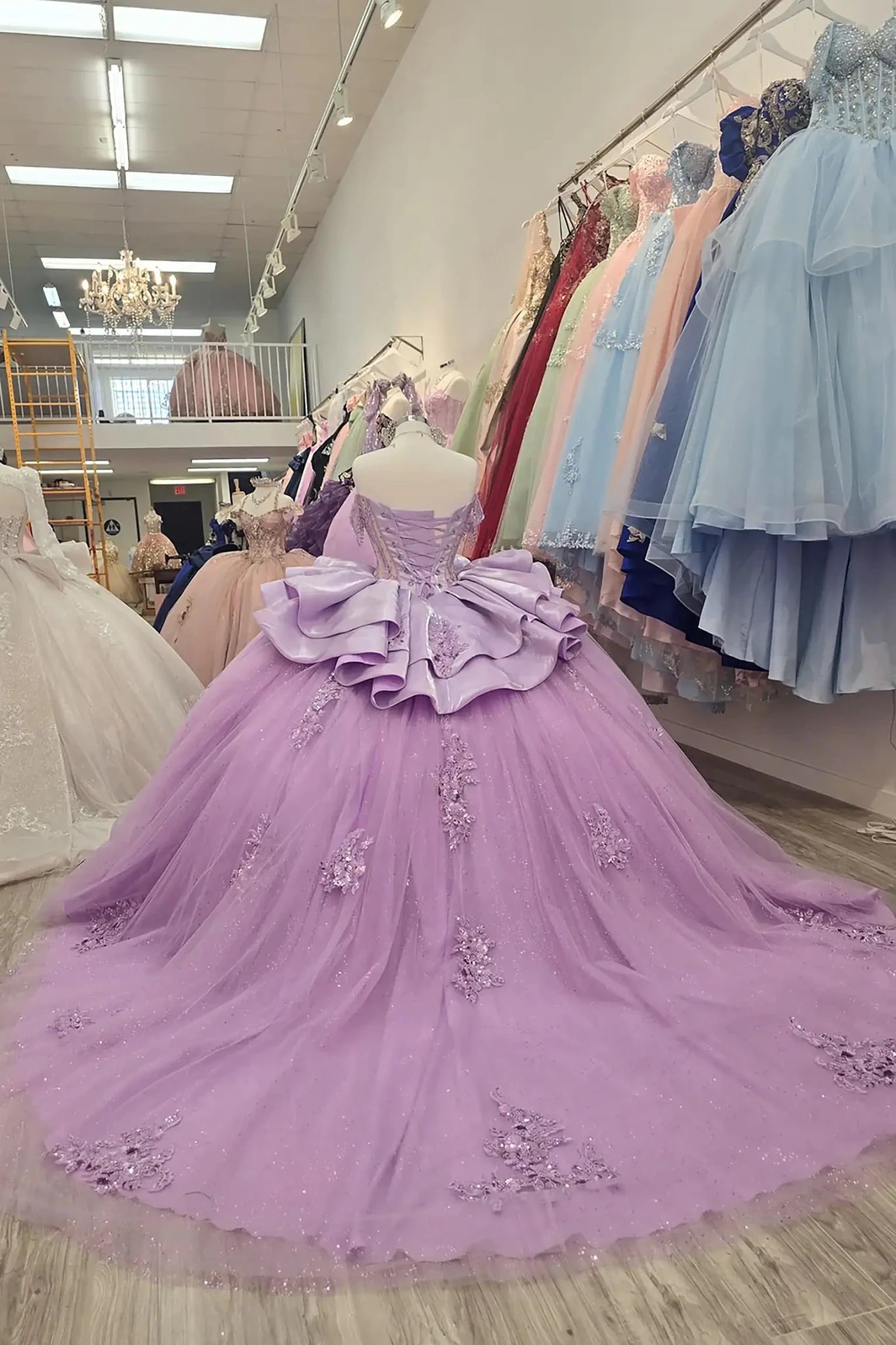 Elegant back full-length the gowns showcase voluminous glitter tulle skirts adorned with intricate floral lace appliqués and tiered, sculptural satin ruffles, displayed in a boutique showroom.