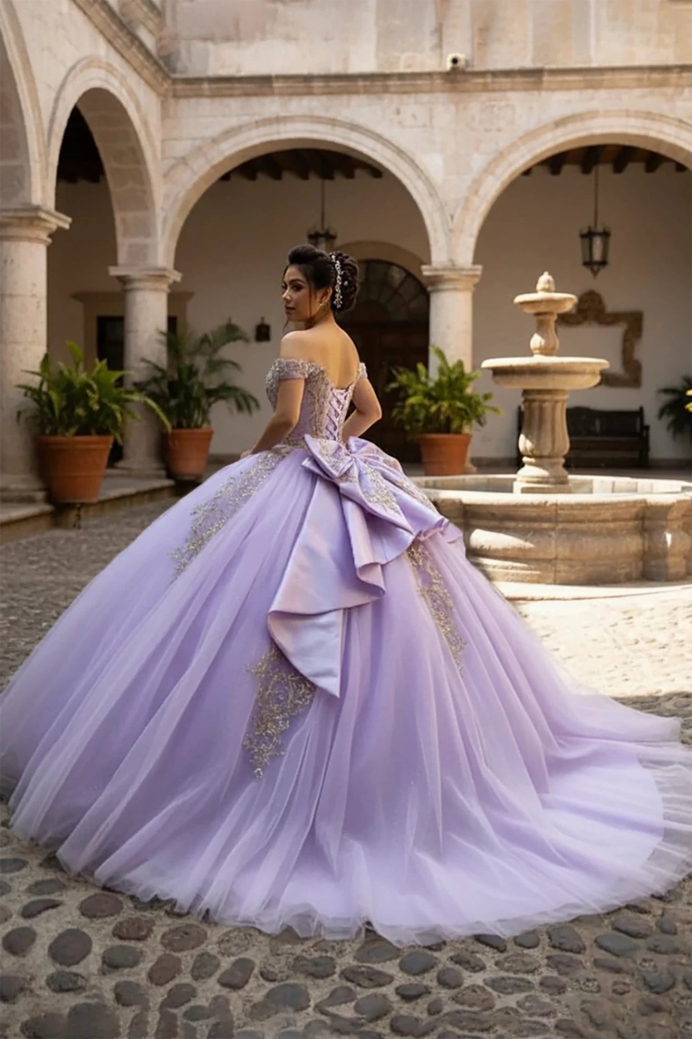Woman in a lilac ball gown with big satin back bow and long train standing in an outdoor courtyard with arches and a fountain.