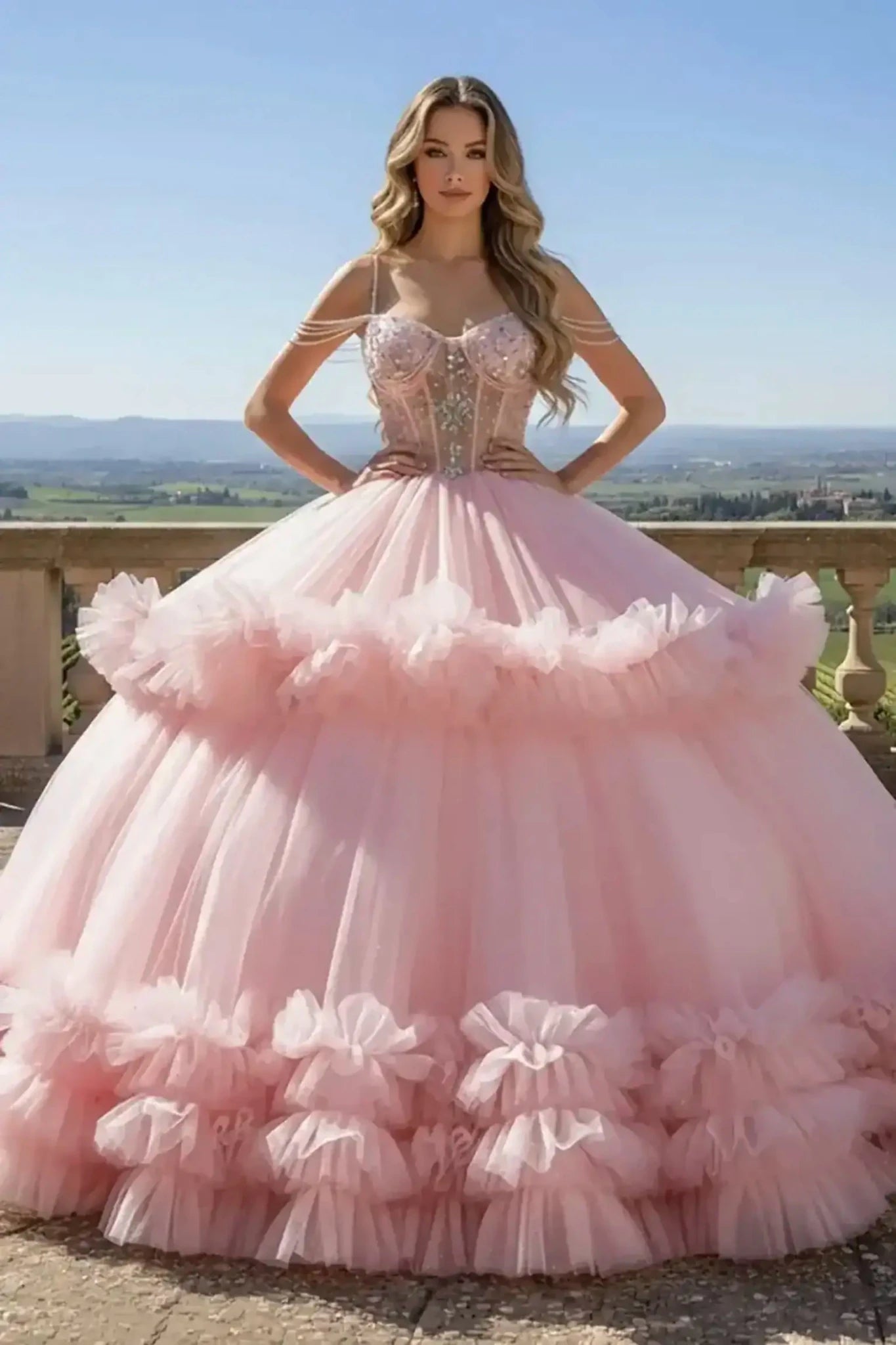 Pink ballgown featuring a beaded corset bodice with pearl shoulder straps and a voluminous tiered ruffled tulle skirt, shown on a model on a scenic stone balcony.