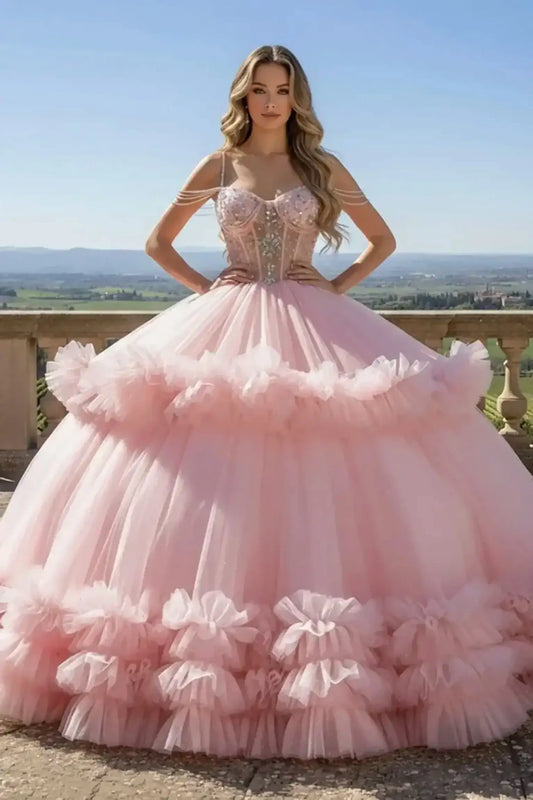 Pink ballgown featuring a beaded corset bodice with pearl shoulder straps and a voluminous tiered ruffled tulle skirt, shown on a model on a scenic stone balcony.