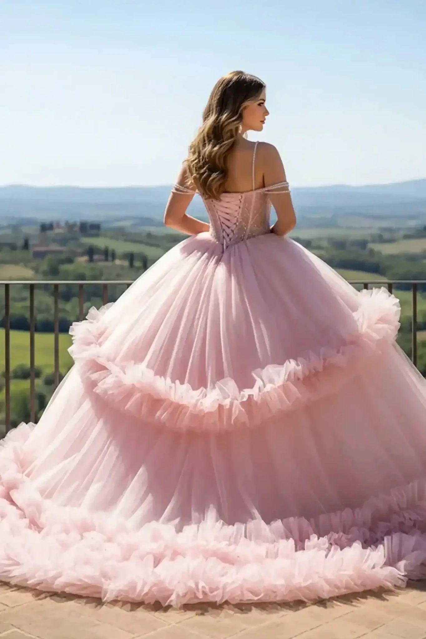 Back view of a Pink ballgown featuring a lace-up corset back, pearl-draped sleeves, and a voluminous tiered ruffled tulle skirt, overlooking a lush vineyard landscape.