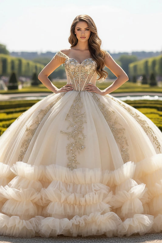 Woman in a luxurious champagne and gold embroidery ball gown with full layered tulle skirt standing in an outdoor setting with greenery.