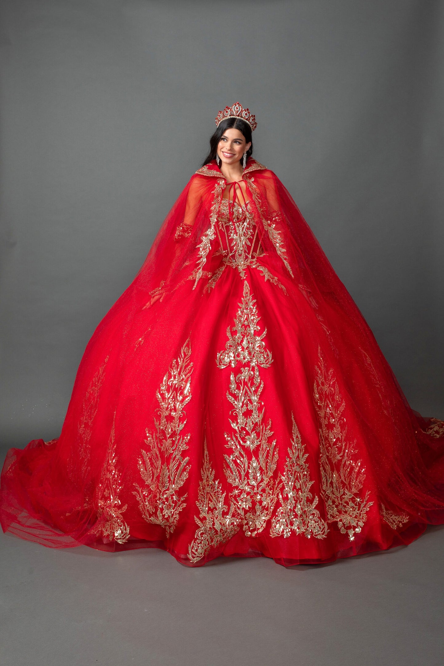 Woman wearing red off-shoulder ball gown quinceañera dress with embroidered corset bodice, ornate appliqué, detachable hooded cape, and full tulle skirt