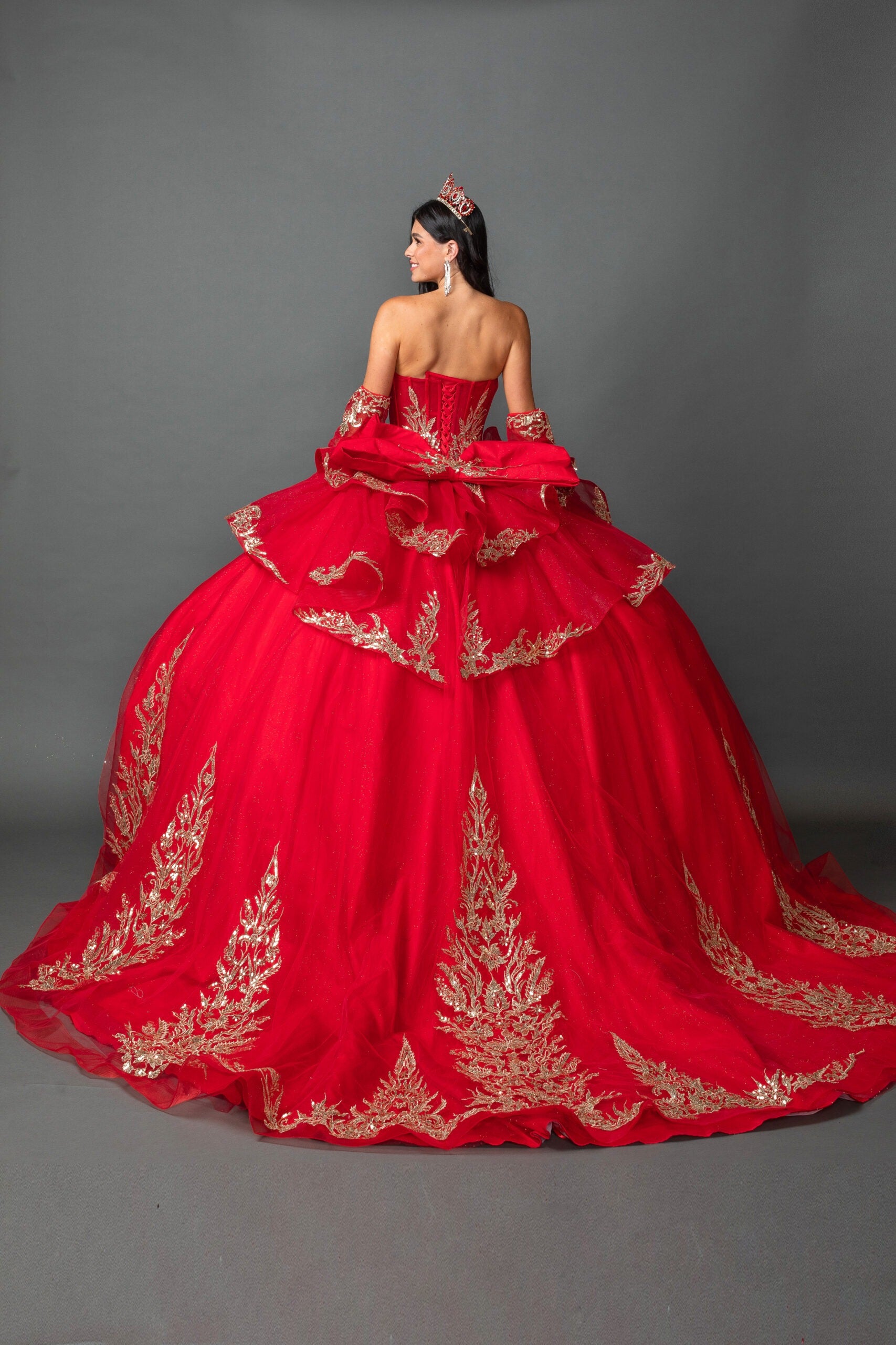 woman back-facing in red off-shoulder ball gown quinceañera dress with lace-up corset bodice, ornate appliqué, and detachable hooded cape detail