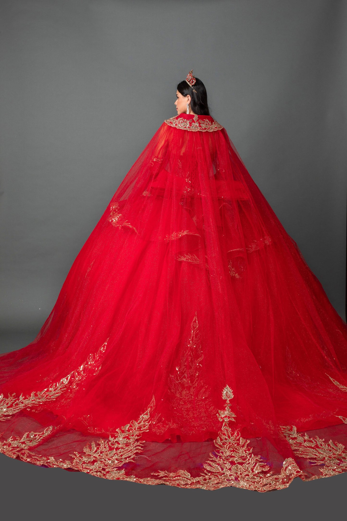 Woman back-facing in red off-shoulder ball gown quinceañera dress with lace-up corset bodice, ornate appliqué, detachable hooded cape, and cascading tulle skirt