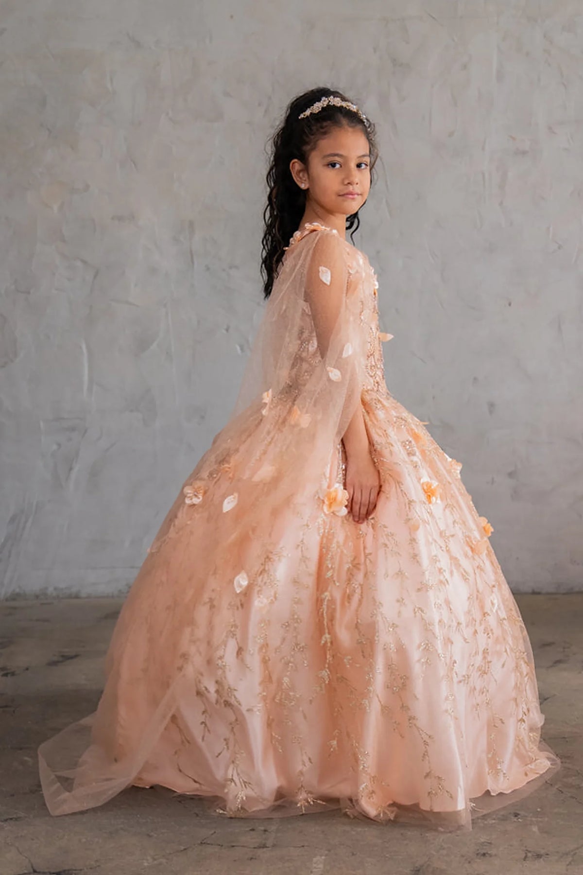At the bodice and skirt, the Blush/Gold mini quince dress showcases off-the-shoulder bodice with dimensional flower trim and removable cape over a glittering tulle skirt.