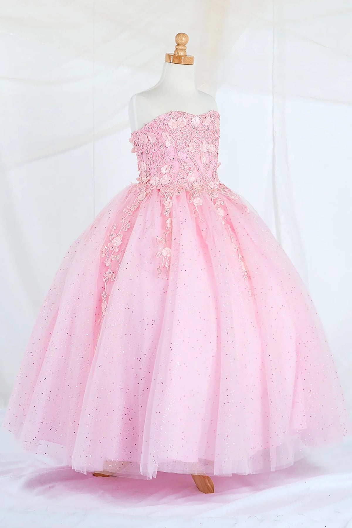 Side-facing display of a bubble-pink mini quince dress featuring a strapless 3D floral appliqué bodice and a full glitter tulle layered skirt.