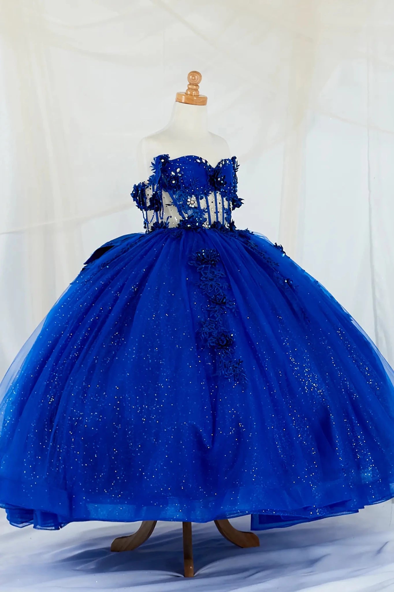 Side-facing display of an elegant royal blue mini quince dress featuring a crystal-beaded bodice, detachable bow accent, and a glitter tulle layered skirt.