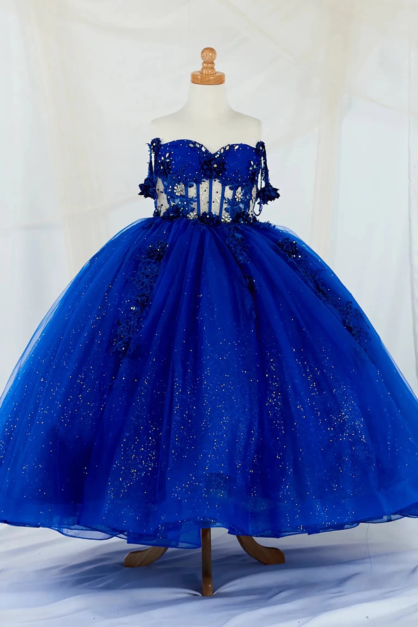 Front-facing display of an elegant royal blue mini quince dress featuring a crystal-beaded bodice, detachable bow accent, and a glitter tulle layered skirt.