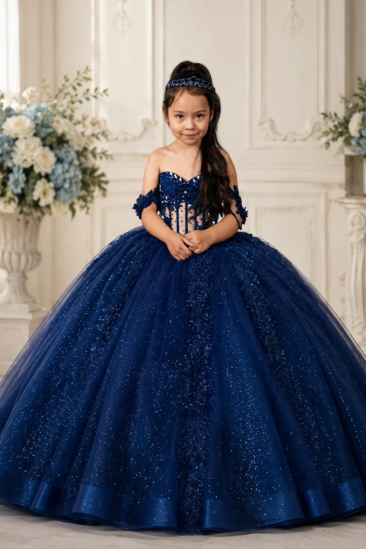 The dress profile reveals, the Navy mini quince dress showcases crystal-beaded bodice with three-dimensional flowers and glitter tulle skirt with a detachable back bow.