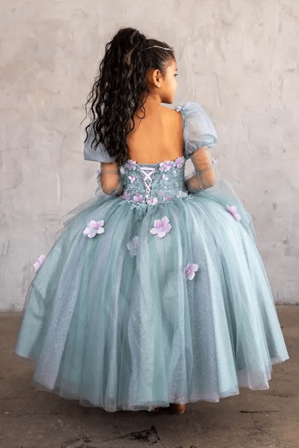 Kid standing back-facing in sage ball gown with lace-up closure, 3D flower detailing, removable sleeves, and cascading skirt